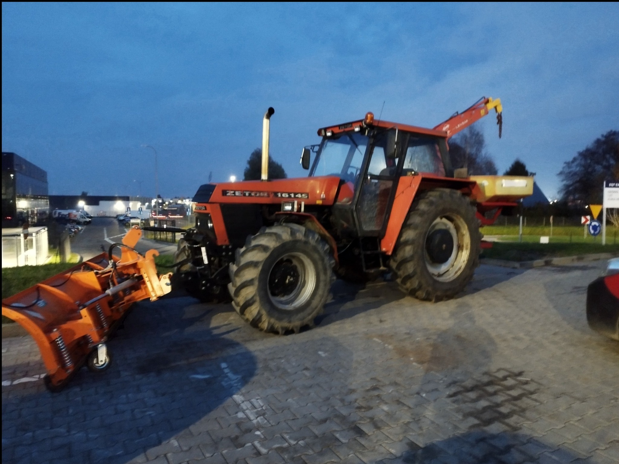 Pomarańczowy traktor Zetor 16145 z pługiem do śniegu na kostce brukowej, gotowy do odśnieżania. Widoczne budynki w tle, zmierzch.