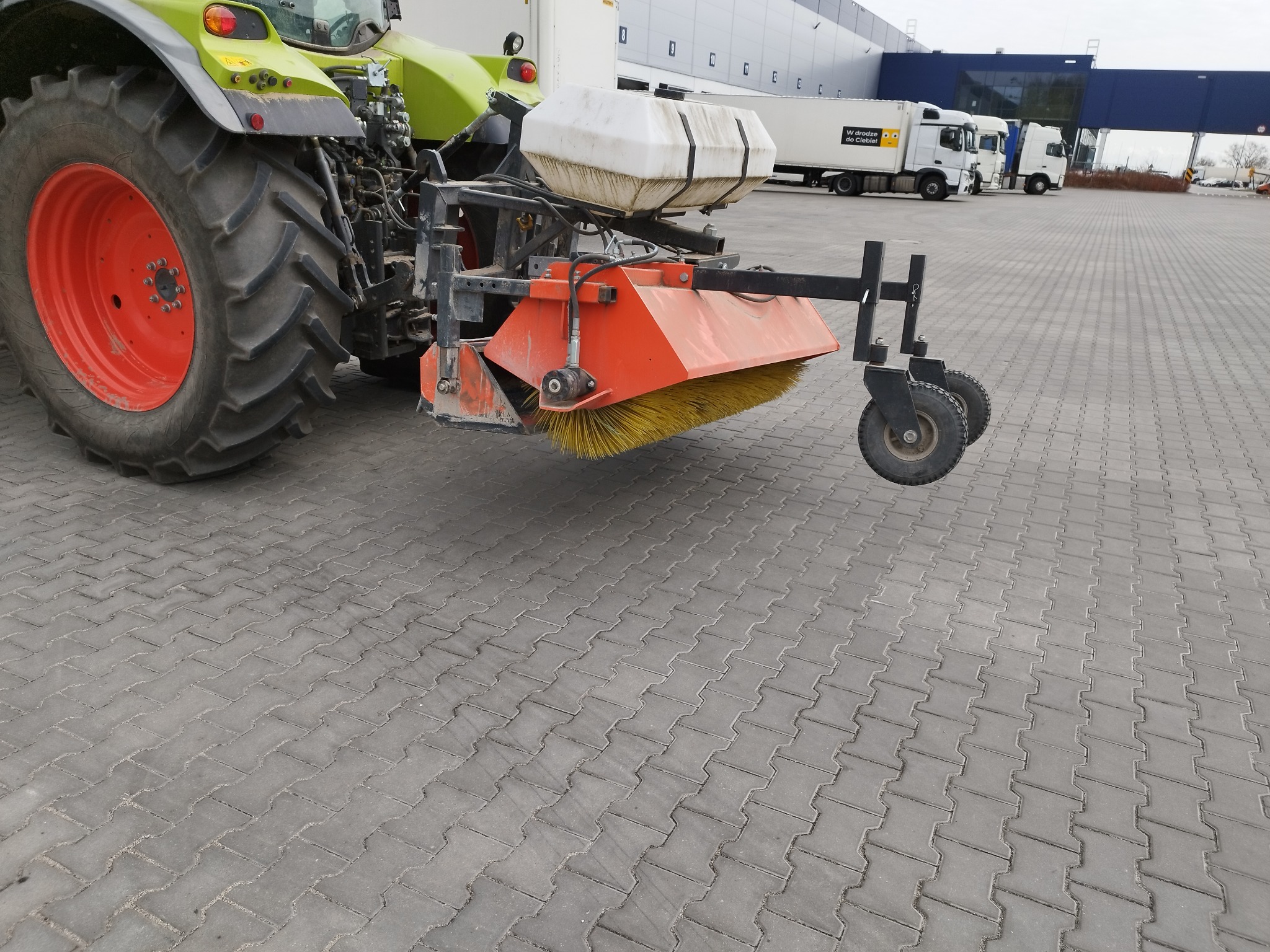 Częściowy widok traktora z zamontowaną szczotką do zamiatania na kostce brukowej. W tle zaparkowane ciężarówki i budynek magazynowy.