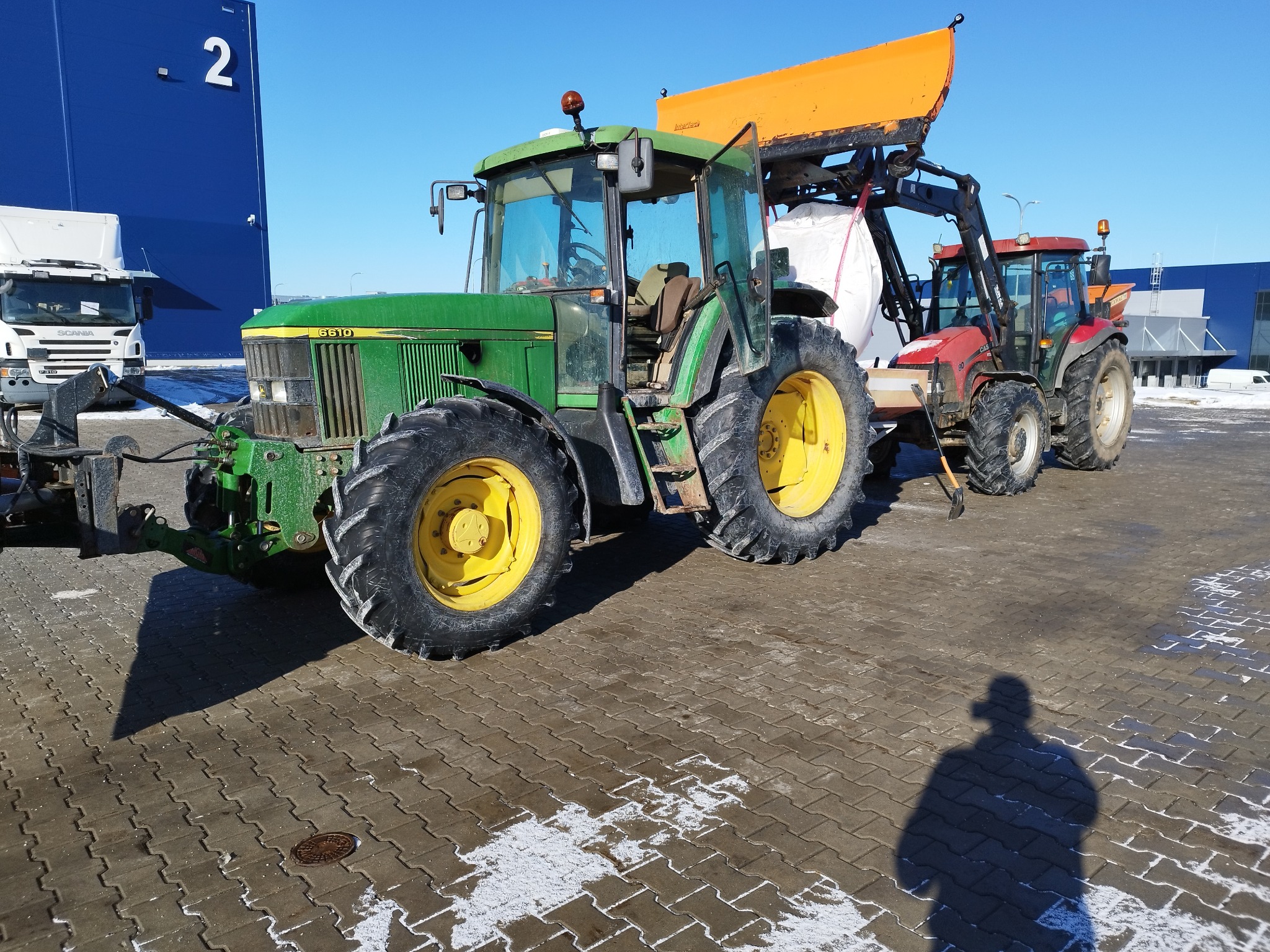 Dwa traktory, zielony John Deere z otwartymi drzwiami i czerwony Case, z zamontowanymi pługami do odśnieżania, stoją na kostce brukowej z resztkami śniegu.