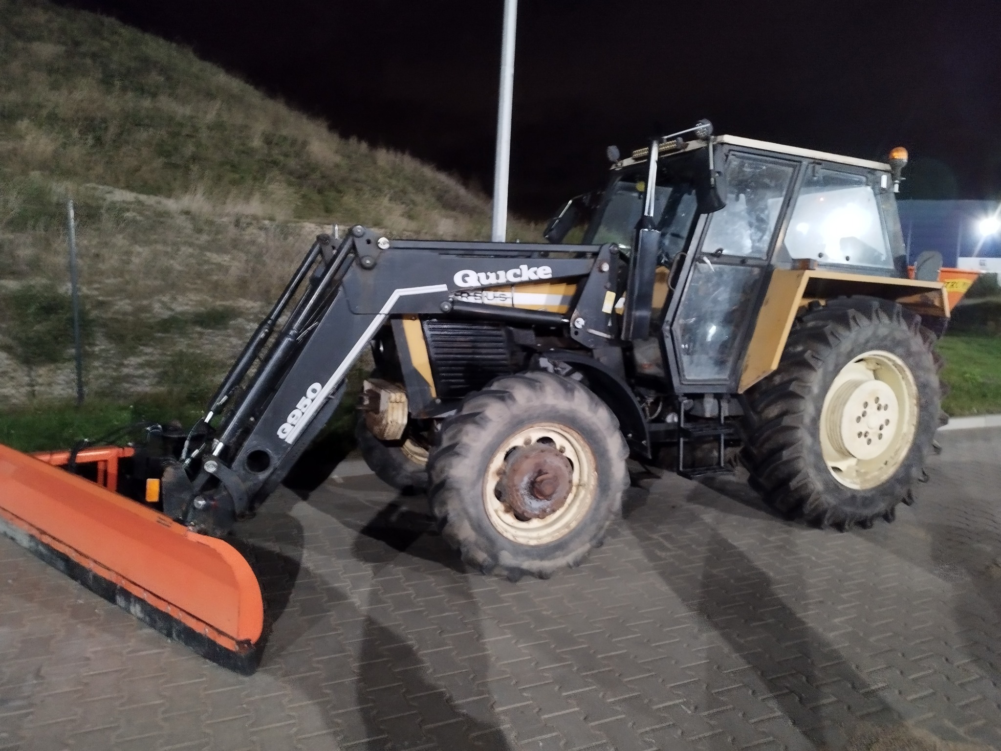 Ciężki traktor z pługiem śnieżnym Quicke 9050, gotowy do pracy na placu. Widoczne oświetlenie nocne i teren pagórkowaty w tle.