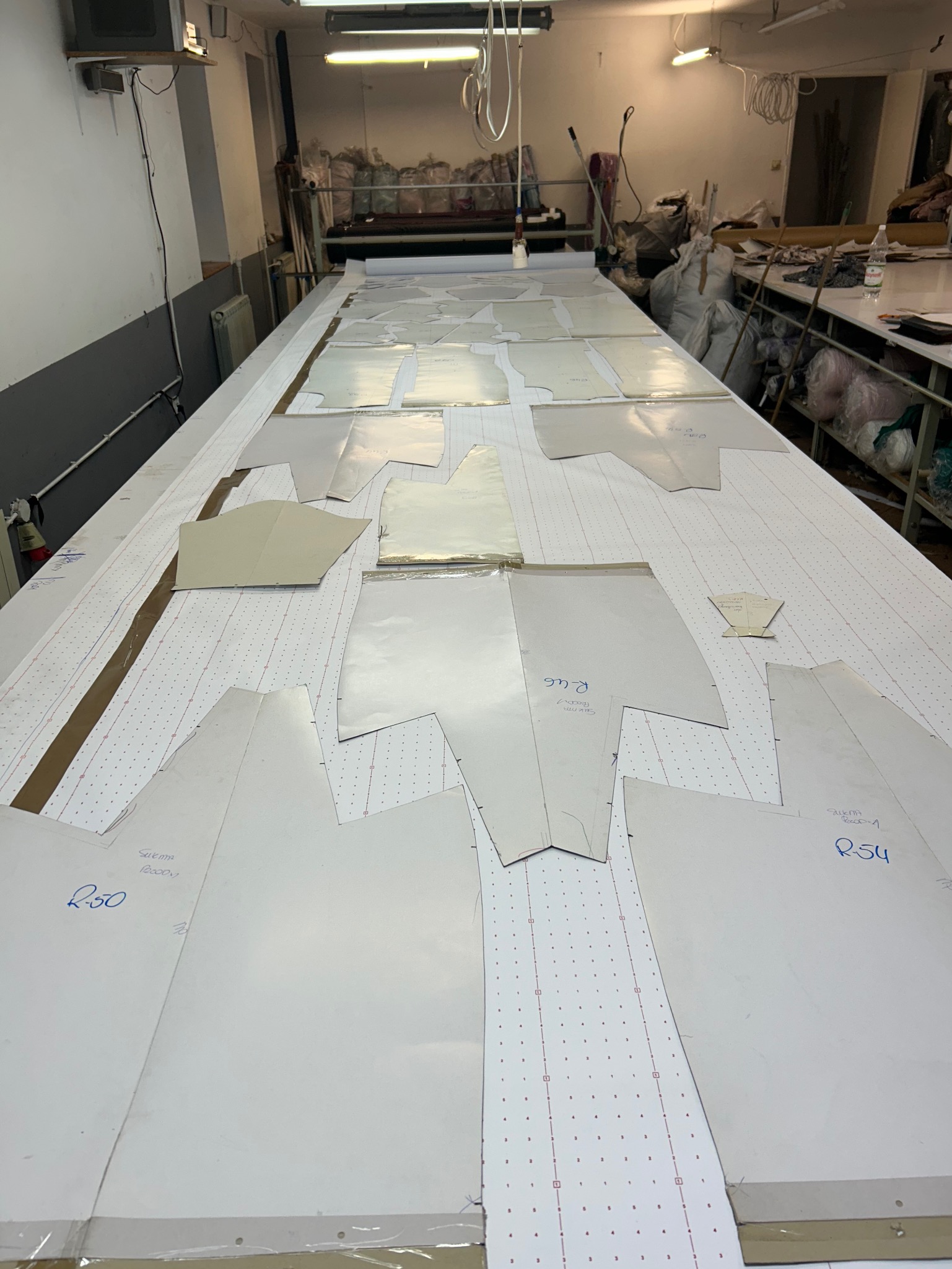 Rozłożone szablony wykrojów odzieży na stole krojowni, przygotowane do cięcia. Widoczne oznaczenia rozmiarów i nazwy elementów garderoby. W tle maszyny i materiały.