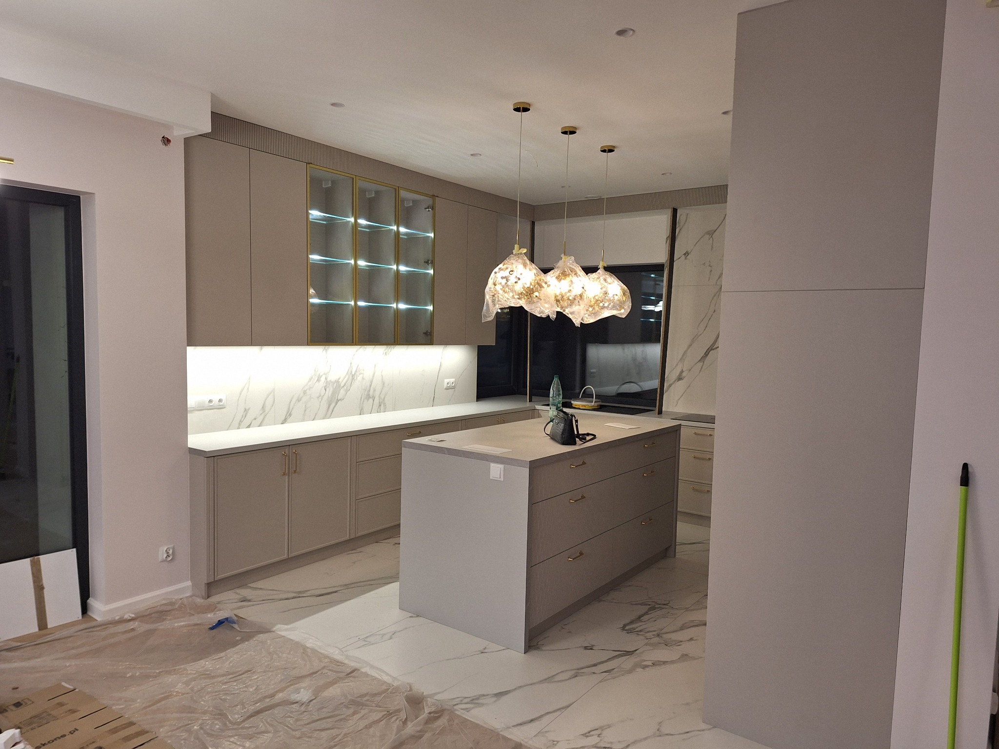 Elegancka kuchnia z wyspą i marmurowym blatem. Szafki w odcieniach beżu z złotymi akcentami. Nad wyspą wiszą trzy ozdobne lampy. Podłoga wyłożona marmurowymi płytami.