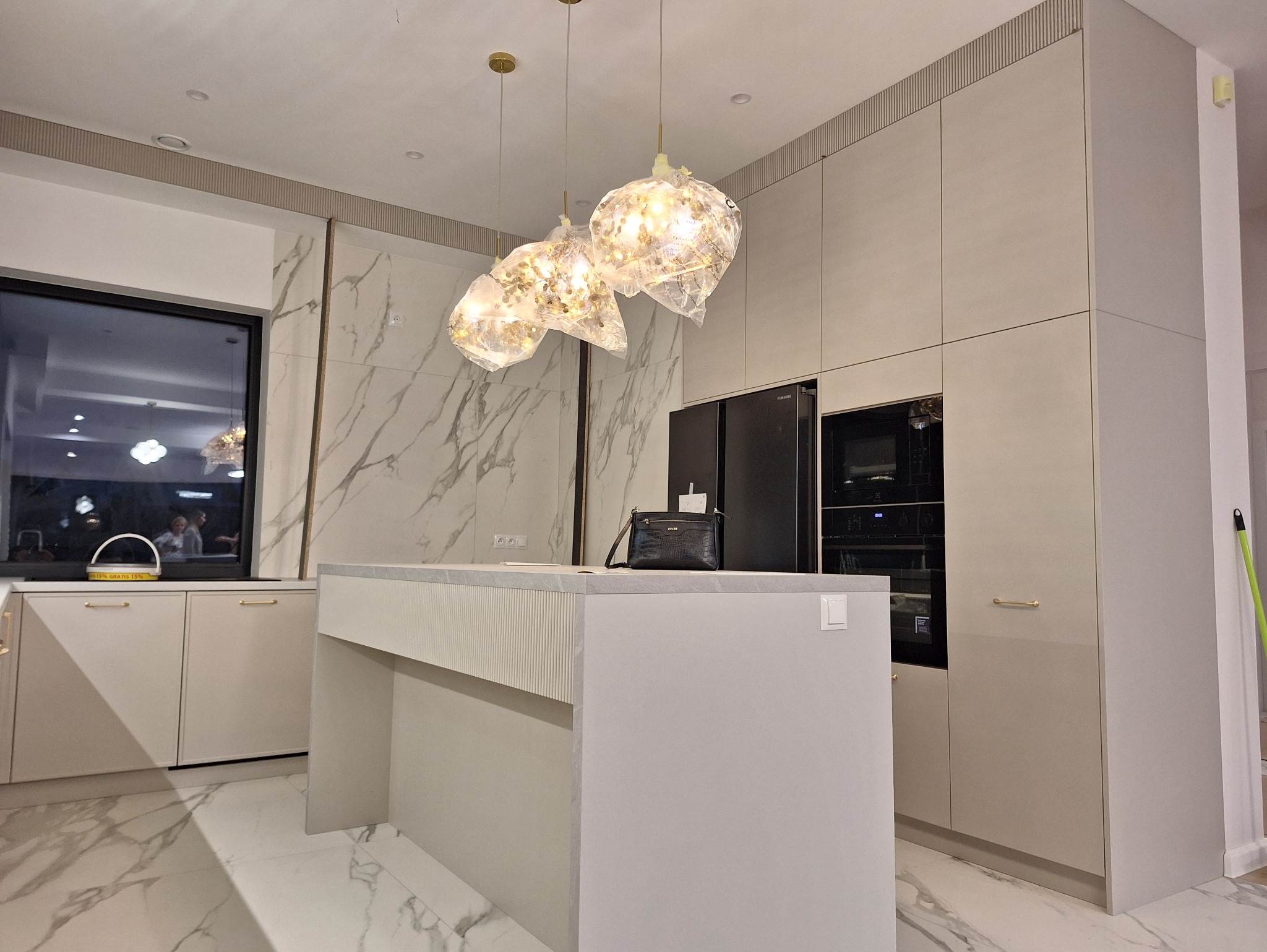 Jasna kuchnia z marmurową ścianą, wyspą i designerskimi lampami. Uwagę przykuwają złote uchwyty szafek i wzór marmuru na podłodze, odbijający światło.