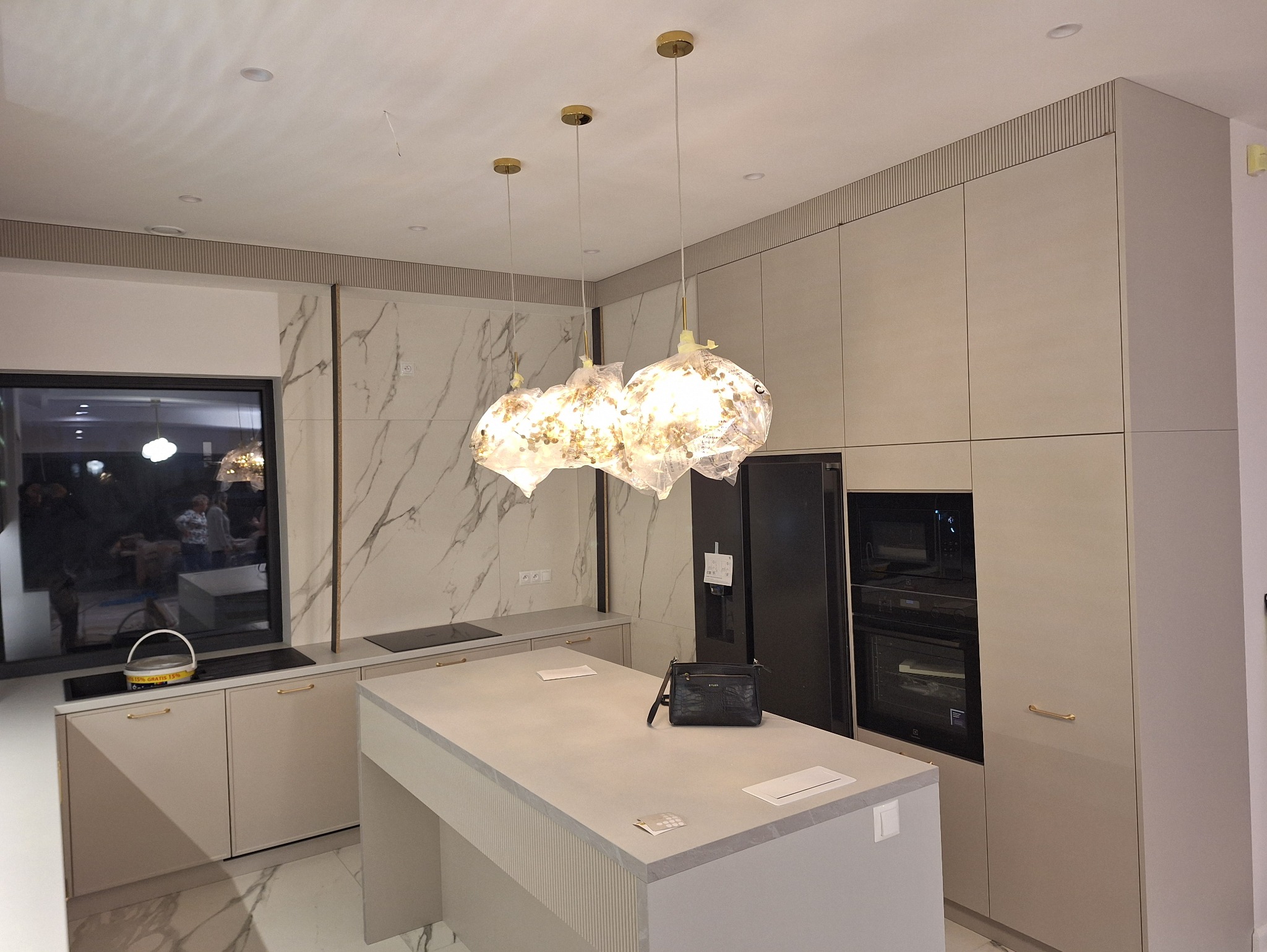 Jasna kuchnia z wyspą i marmurową ścianą. Nad wyspą wiszą trzy oryginalne lampy. Na blacie leży czarna torebka. Widoczne zabudowane sprzęty AGD.