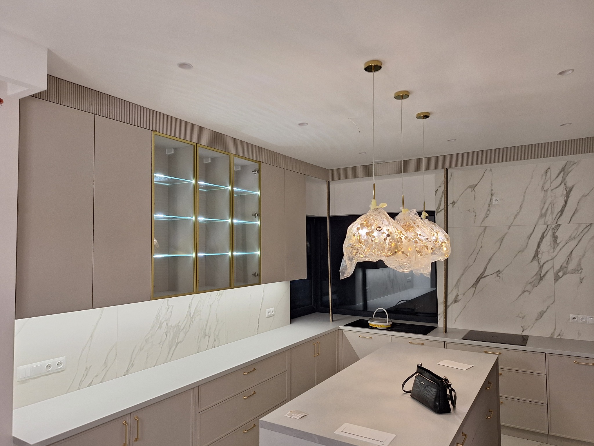 Elegancka kuchnia z beżowymi szafkami, złotymi akcentami i marmurowym backsplashiem. Nad wyspą wiszą ozdobne lampy w folii ochronnej. Na blacie leży czarna torebka.