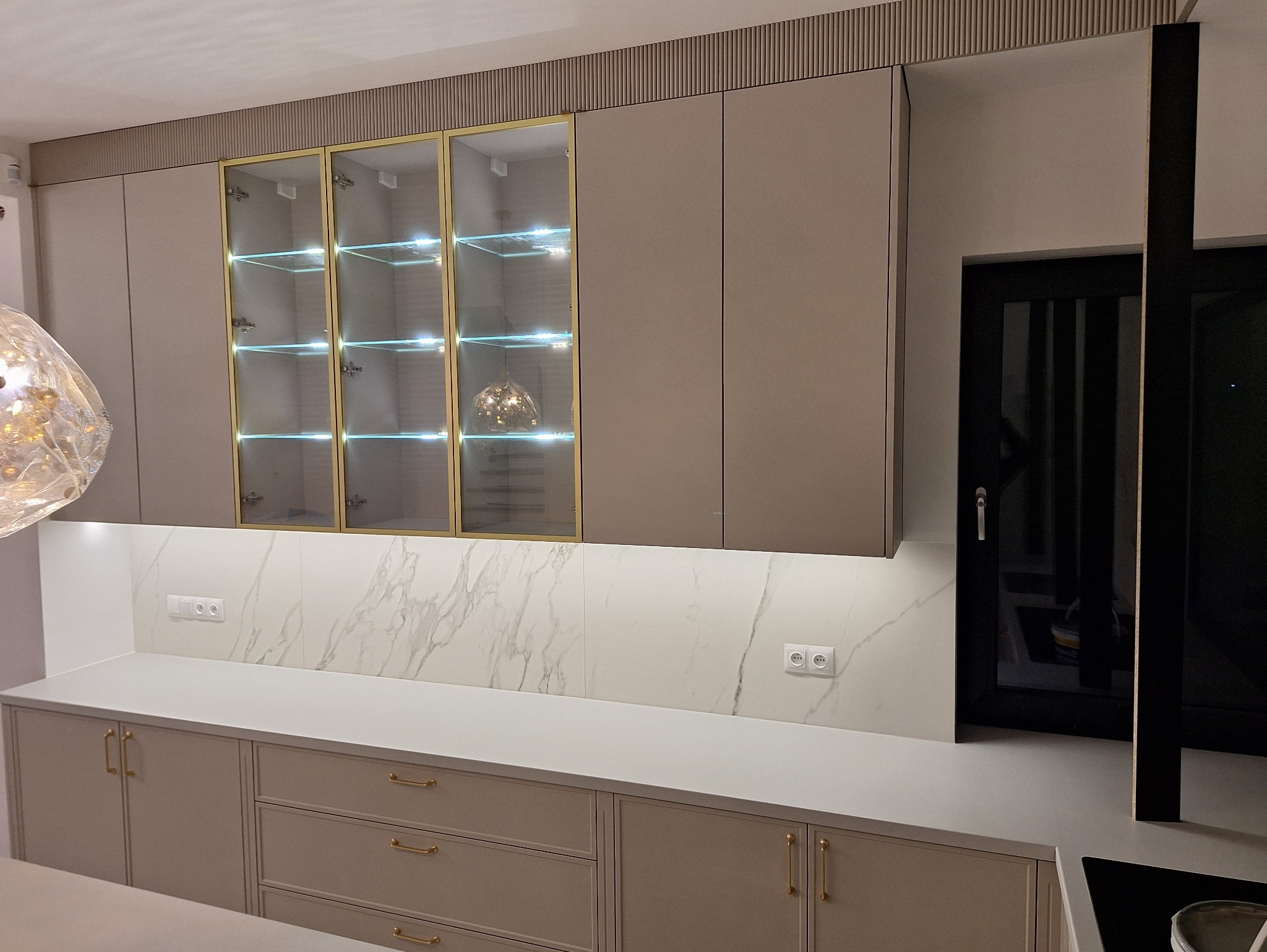 Beżowa kuchnia z przeszklonymi szafkami górnymi, podświetlonymi półkami i blatem z marmurowym wzorem. Złote uchwyty i listwy dekoracyjne dodają elegancji.