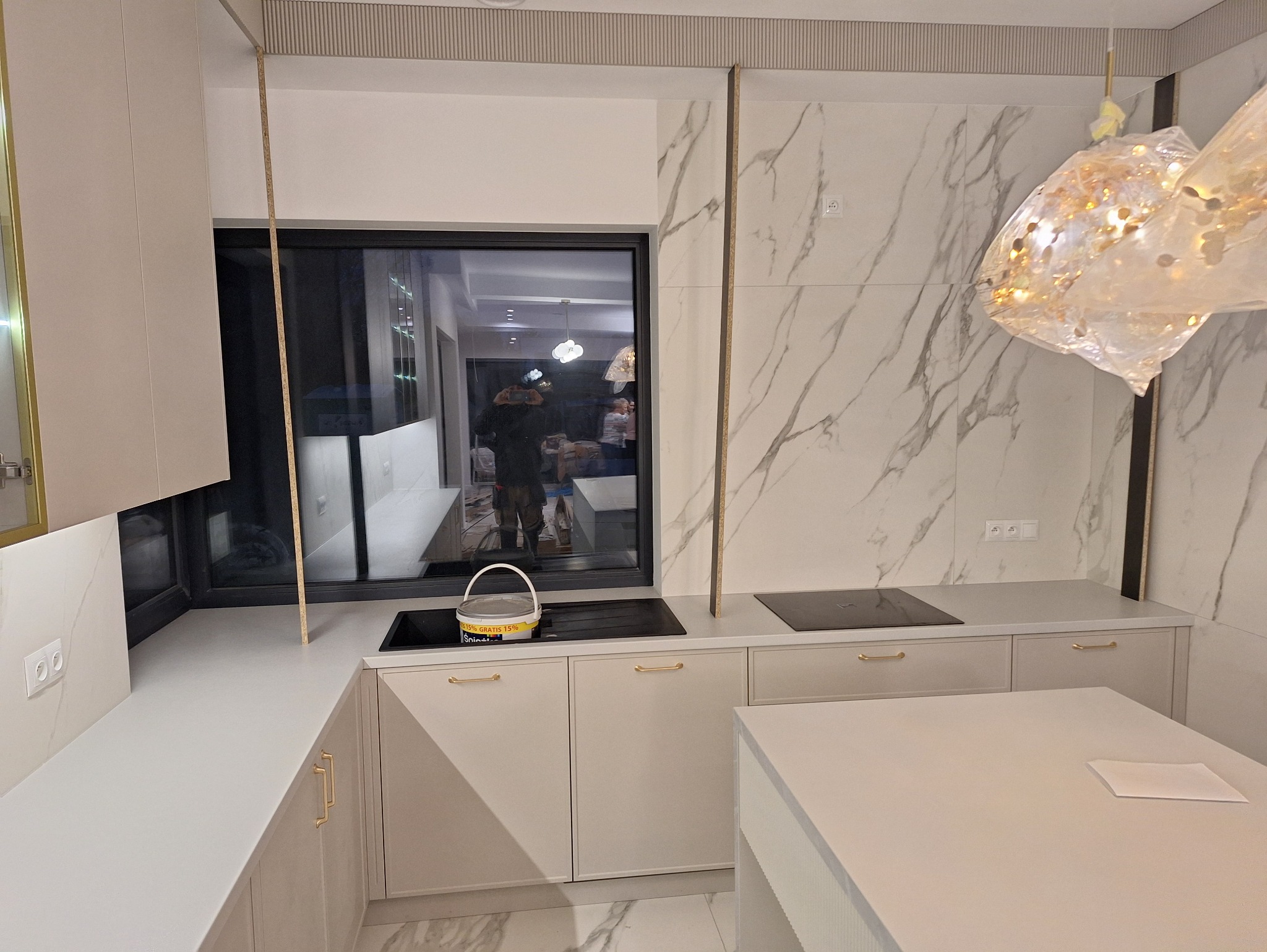 Jasna kuchnia na wymiar z marmurowym akcentem w Lublinie. Widoczny fragment wyspy kuchennej, płyta grzewcza i dekoracyjna lampa. W oknie odbicie wnętrza.