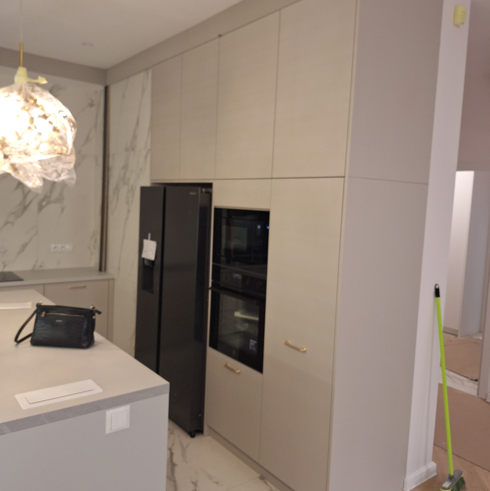 Jasna kuchnia z zabudową: lodówka, piekarnik i szafki w jednej linii. Na blacie torebka i gniazdko. Marmurowe wykończenie ściany i designerska lampa.