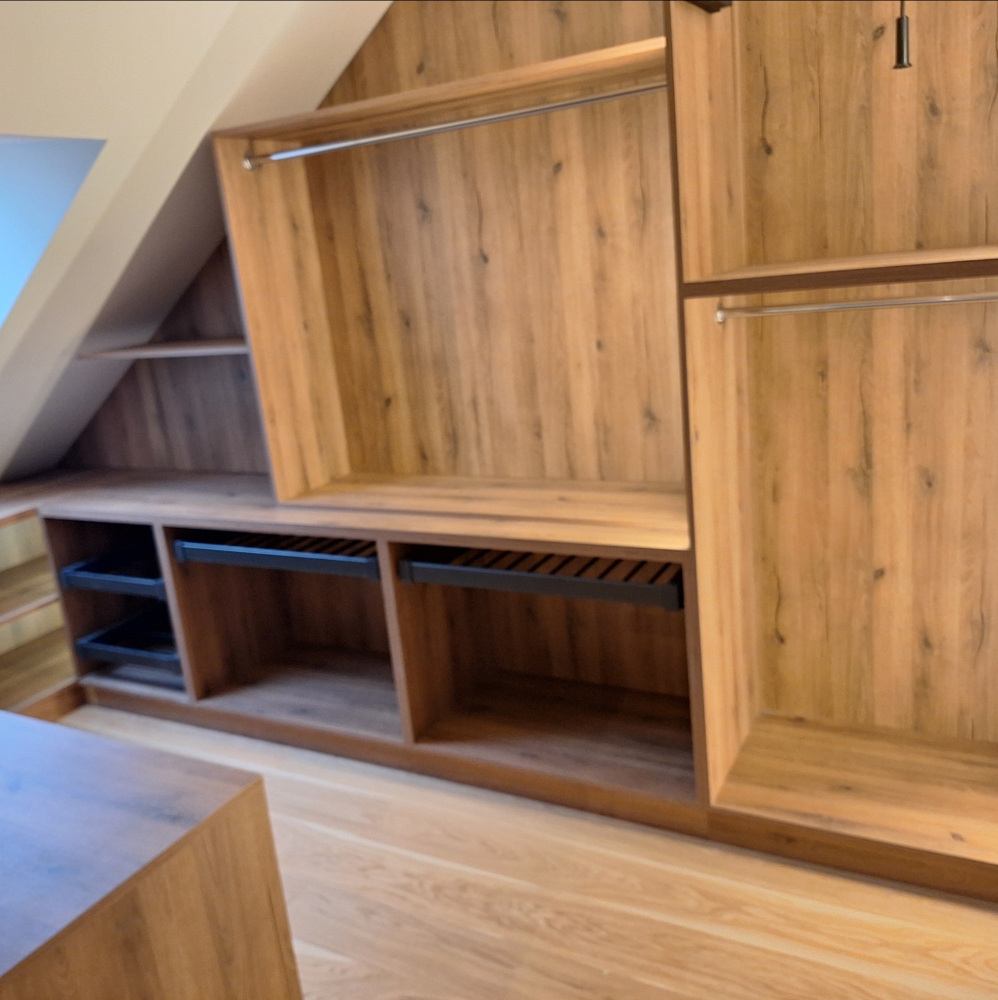 Drewniana szafa wnękowa z półkami i drążkami, dopasowana do skosu dachu. Widoczne kosze i szuflady. Wykończenie w ciepłym odcieniu drewna.