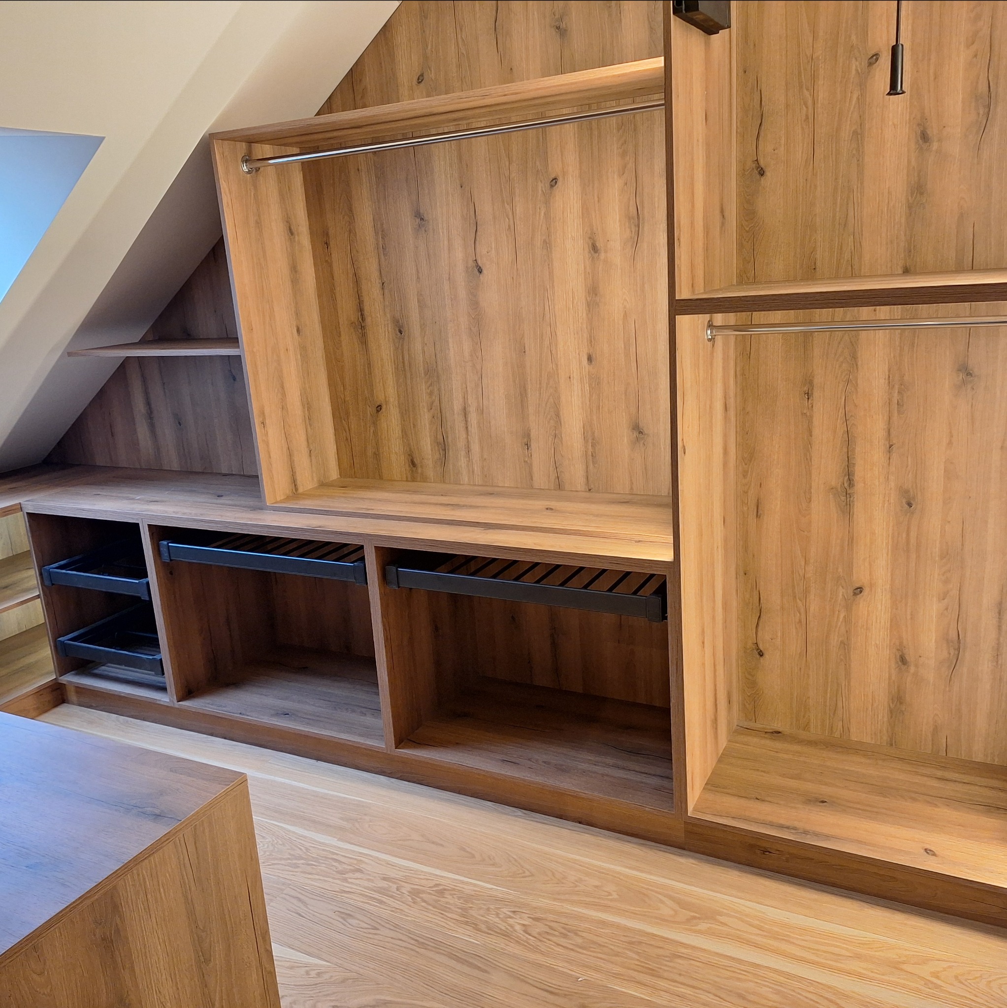 Drewniana zabudowa garderoby na poddaszu ze skośnym sufitem, z wysuwanymi koszami i drążkami na ubrania. Widoczna struktura drewna i metalowe elementy.