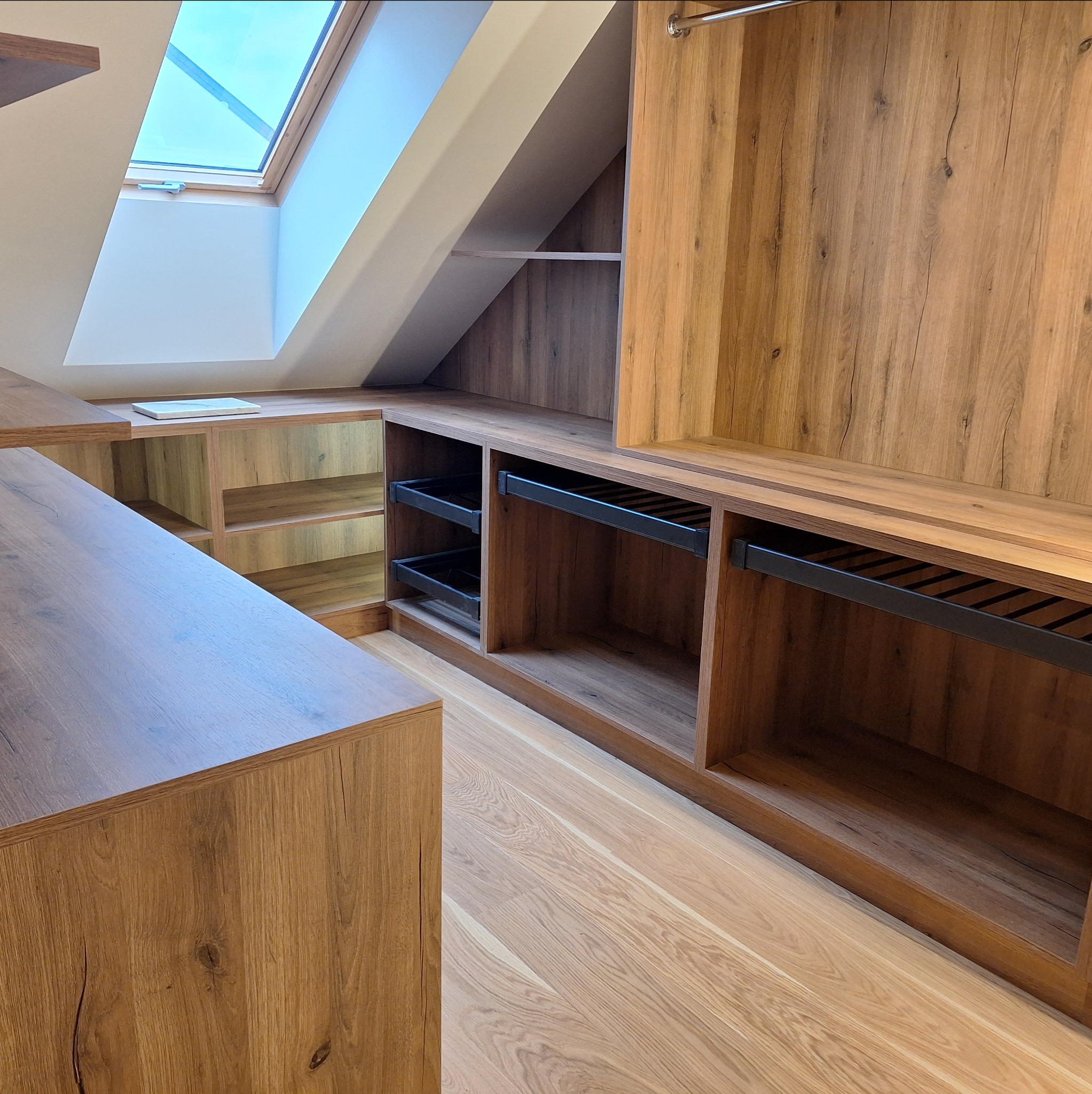 Drewniana zabudowa garderoby na poddaszu ze skosami, z otwartymi półkami i szufladami. Widoczny fragment podłogi z jasnego drewna i okno dachowe.
