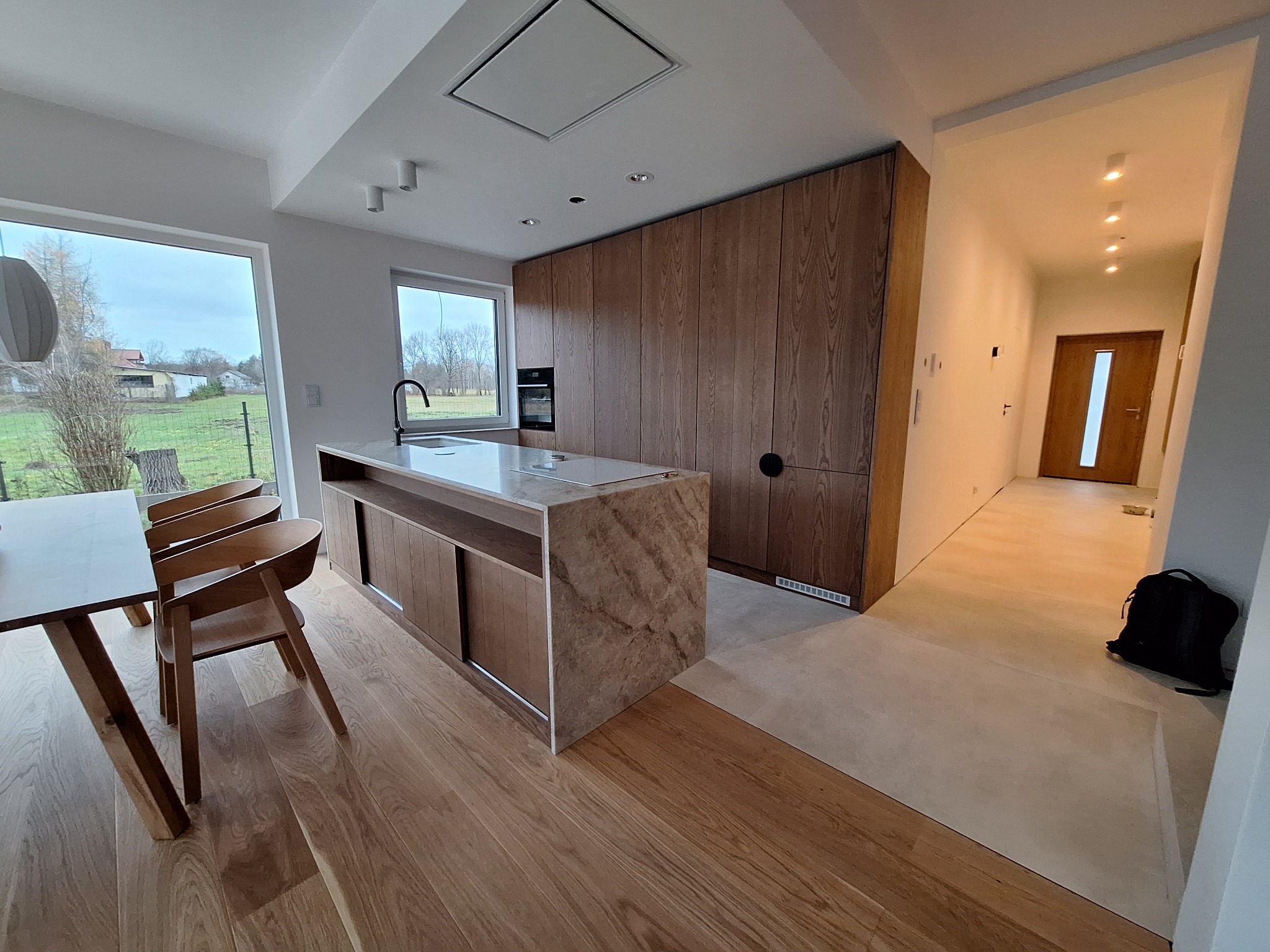 Nowoczesna kuchnia z drewnianą zabudową i wyspą z marmurowym blatem, widok na stół z krzesłami i okno z widokiem na zewnątrz. Minimalistyczny design.