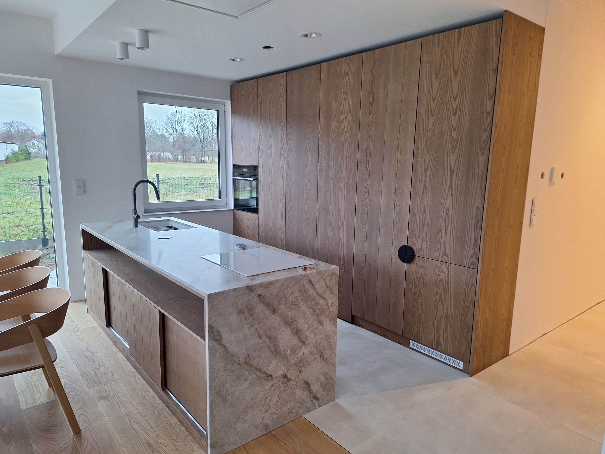 Nowoczesna kuchnia z wyspą o marmurowym wykończeniu i drewnianymi szafkami, z widokiem na zieleń za oknem. Minimalistyczny design, jasne wnętrze.