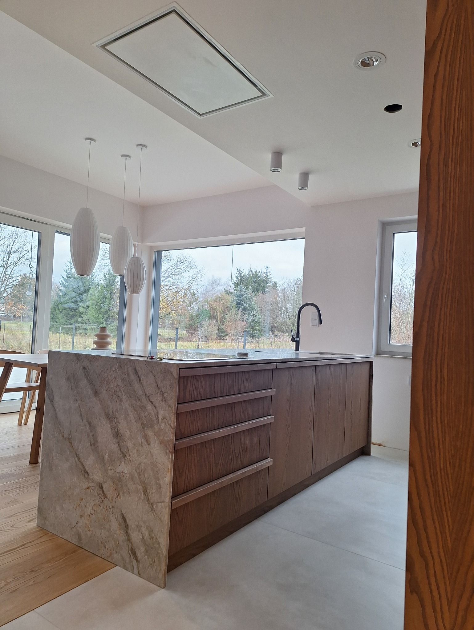 Nowoczesna kuchnia z wyspą wykończoną marmurem, drewniane fronty szafek, czarna bateria, minimalistyczne lampy wiszące i duże okna z widokiem na zieleń.