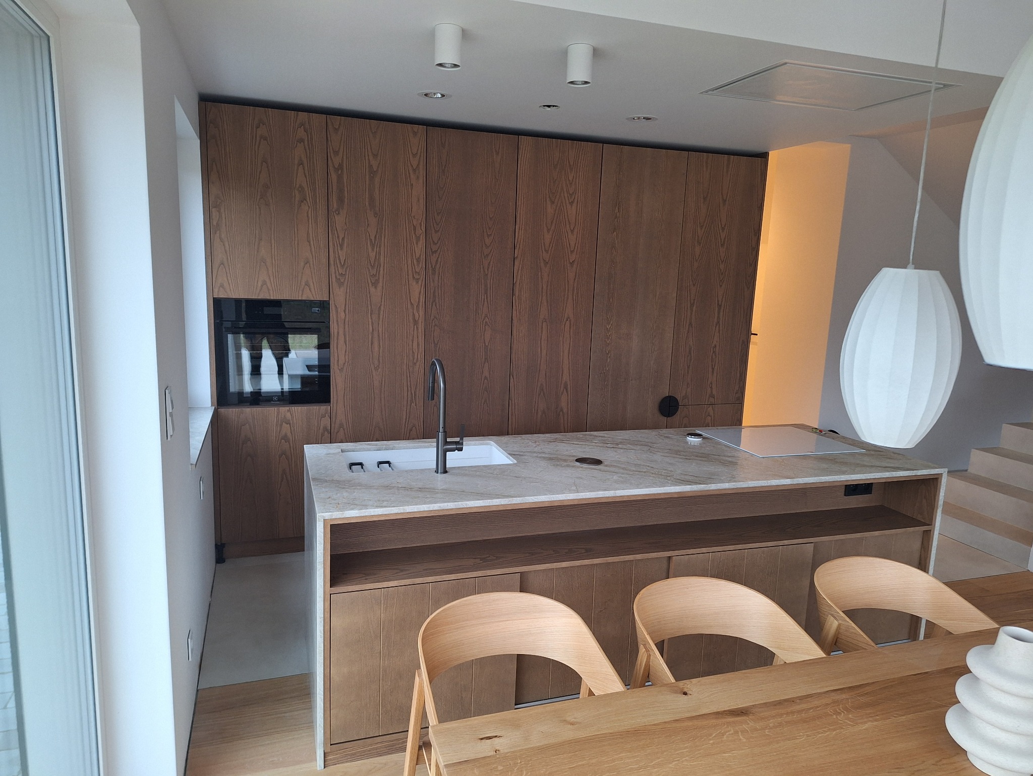 Nowoczesna kuchnia z drewnianymi frontami, marmurowym blatem i wyspą. Minimalistyczne oświetlenie i krzesła z jasnego drewna. Widoczny piekarnik w zabudowie.