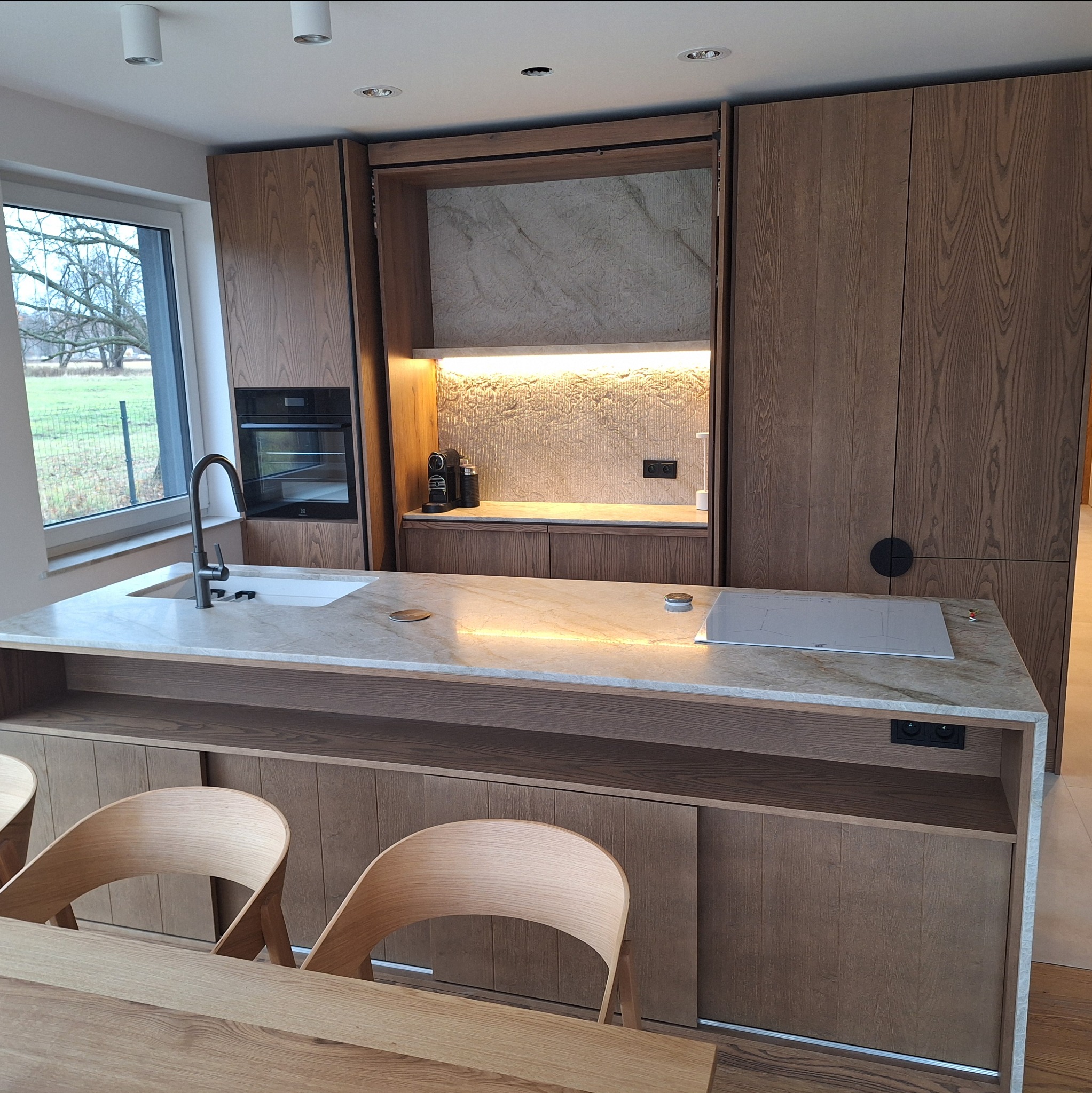 Nowoczesna kuchnia z drewnianymi frontami, marmurowym blatem i wyspą z płytą indukcyjną. Widoczne krzesła i okno z widokiem na zewnątrz. Oświetlenie LED podkreśla detale.