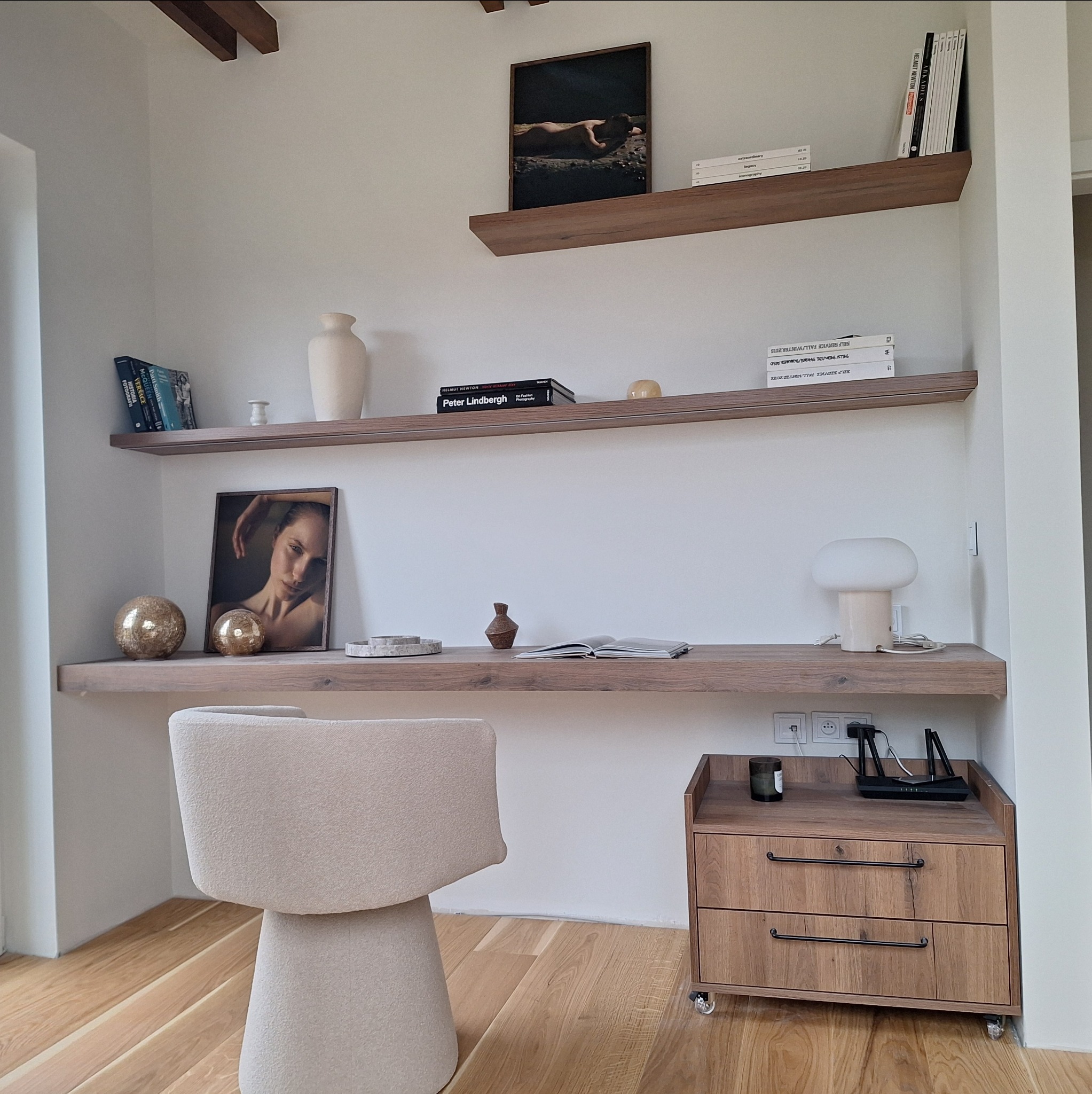 Minimalistyczne biurko z drewnianymi półkami, dekoracjami i krzesłem o nietypowym kształcie na tle jasnej ściany i drewnianej podłogi. Widoczne książki i obrazy.