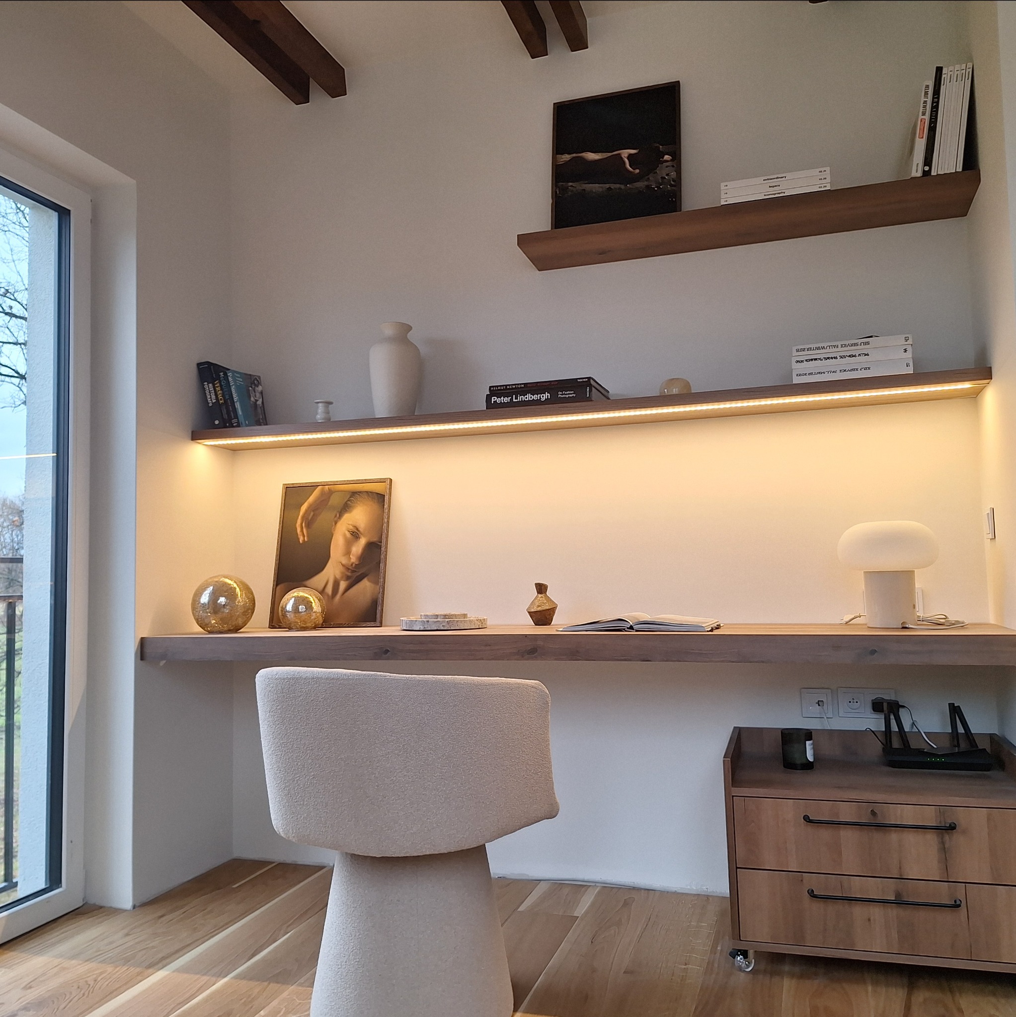 Drewniany blat biurka z oświetleniem LED, dekoracjami i książkami, uzupełniony szafką na kółkach. Jasne wnętrze z drewnianą podłogą i elementami dekoracyjnymi.