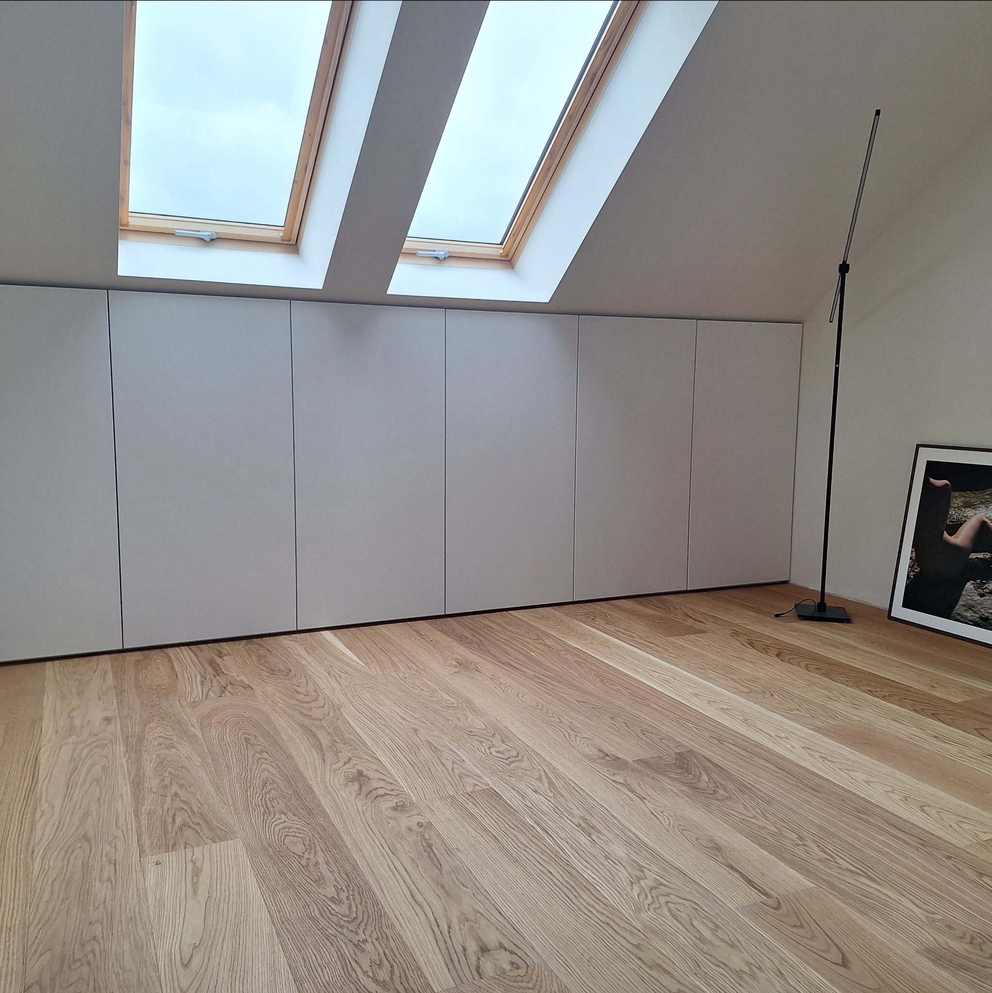 Biała szafa wnękowa pod skosami dachu z oknami, na drewnianej podłodze. Minimalistyczny design, oświetlenie lampą podłogową i obrazem w ramie.
