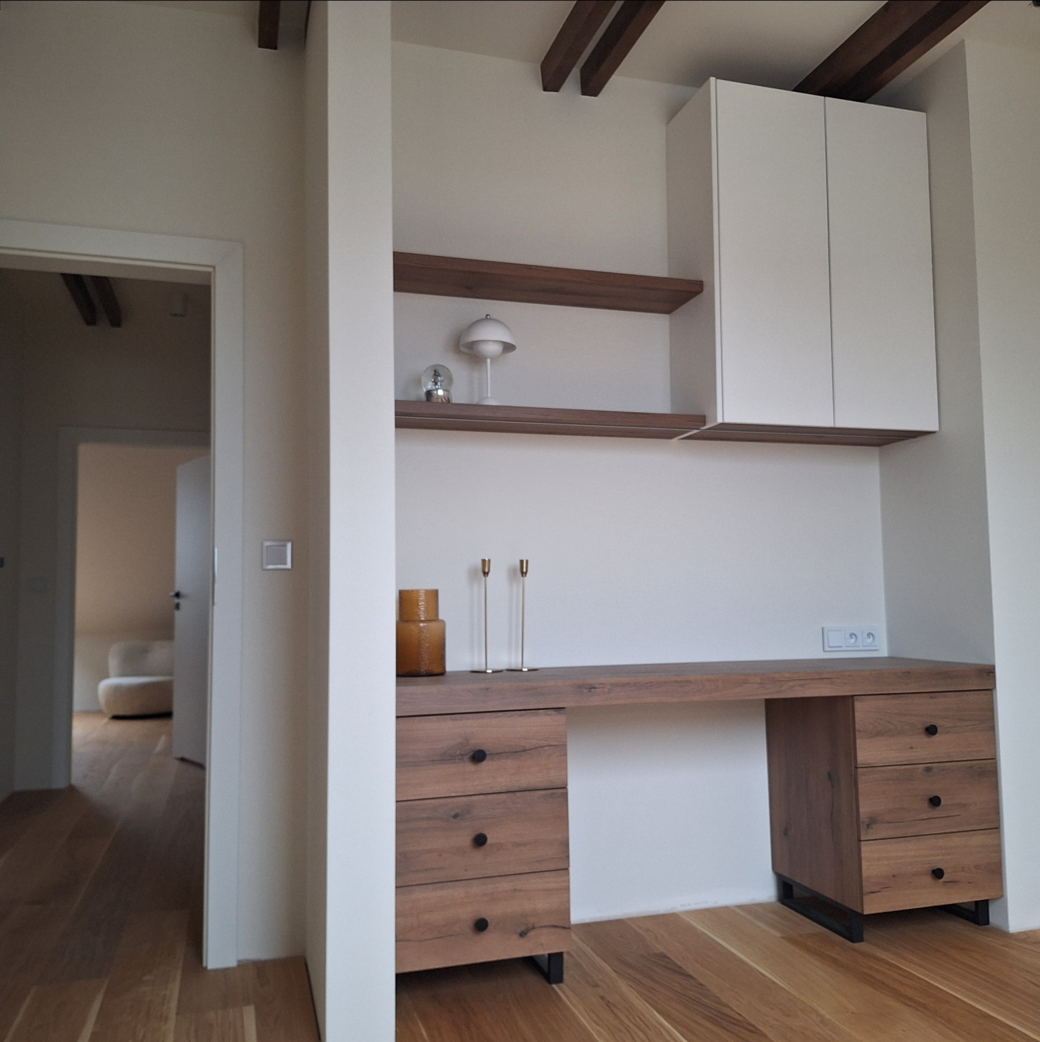 Drewniane biurko z szufladami i szafką wiszącą w jasnym wnętrzu, uzupełnione półkami i dekoracjami. Widok z perspektywy wejścia do pokoju.