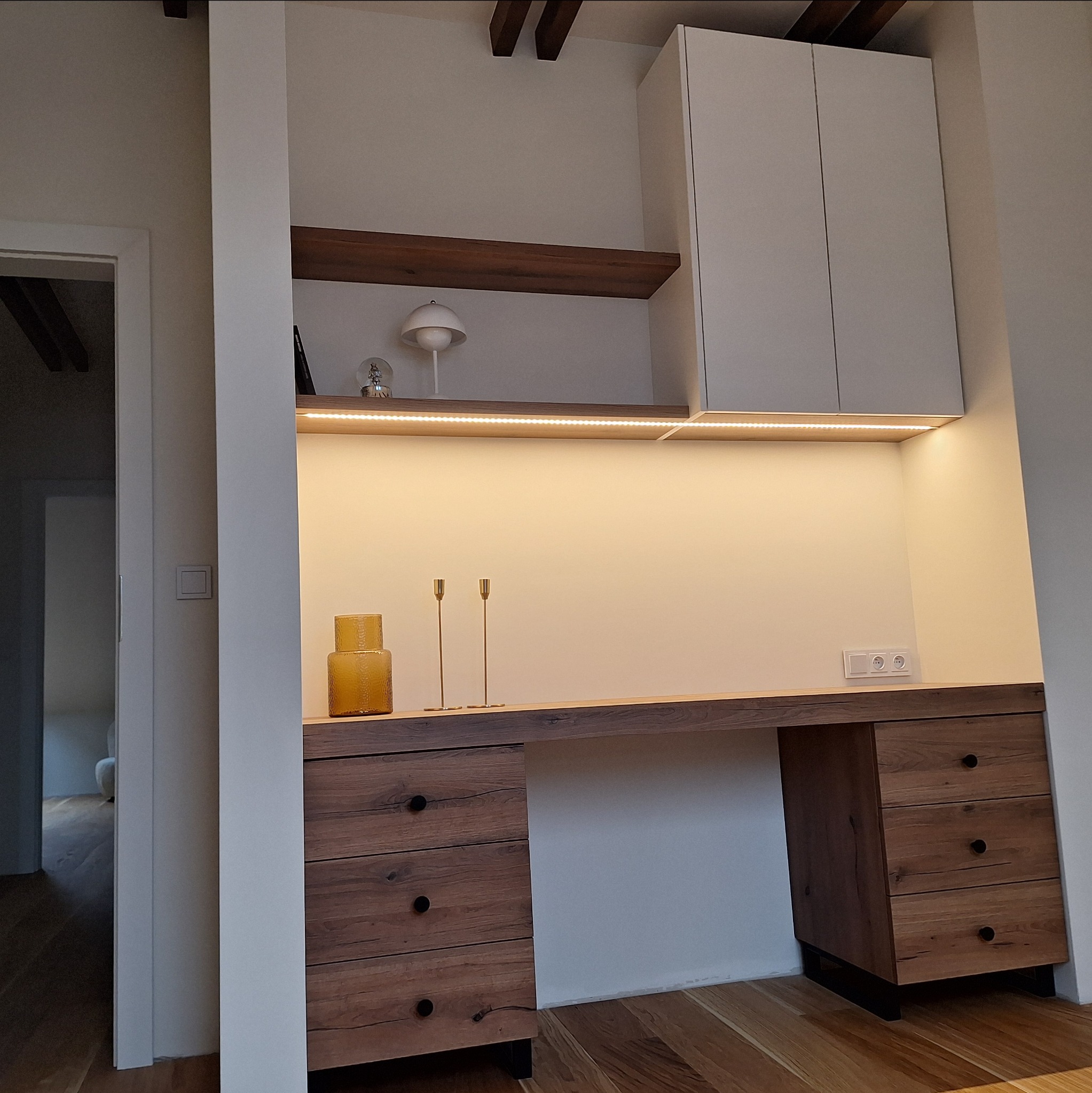 Drewniane biurko z szufladami i półkami podświetlonymi LED, wbudowane w białą zabudowę. Widoczne elementy dekoracyjne: wazon i świeczniki. Minimalistyczny design.