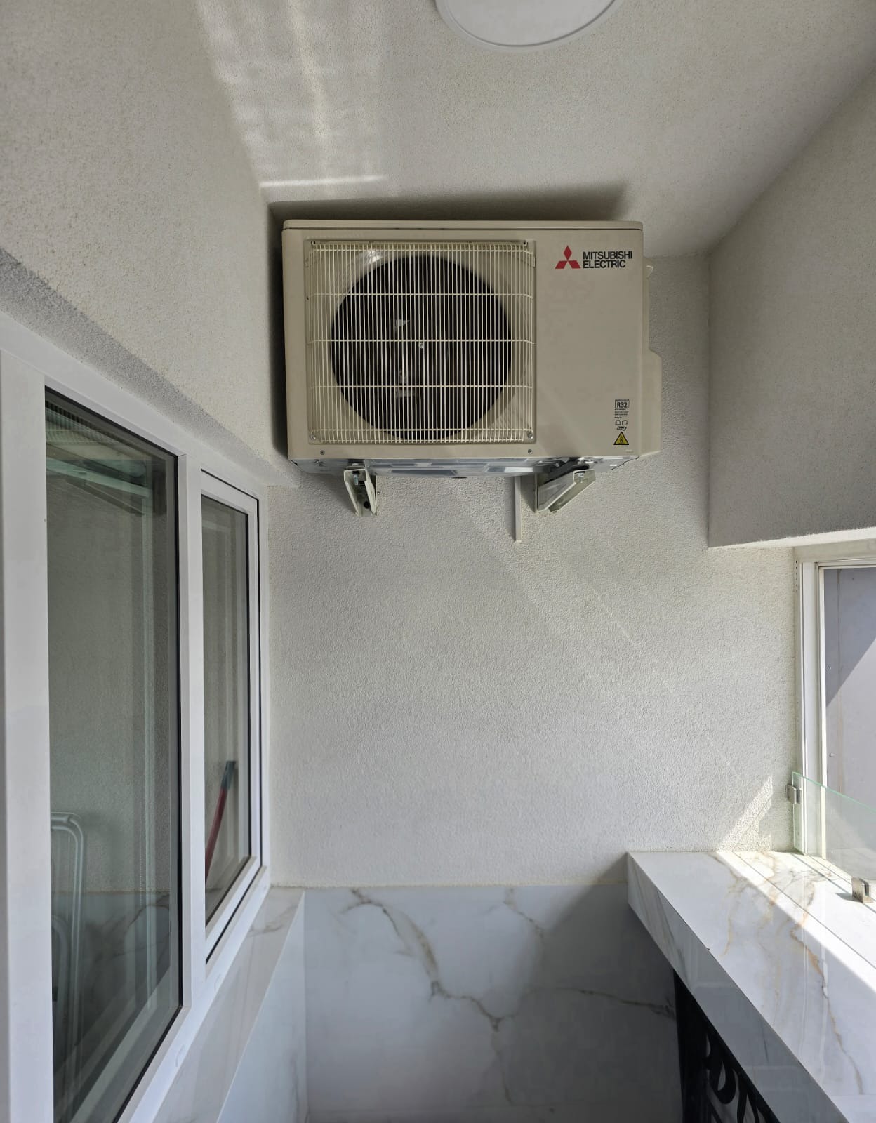 Klimatyzator Mitsubishi Electric zamontowany na ścianie balkonu z marmurowym wykończeniem, widoczny przez okno fragment otoczenia.