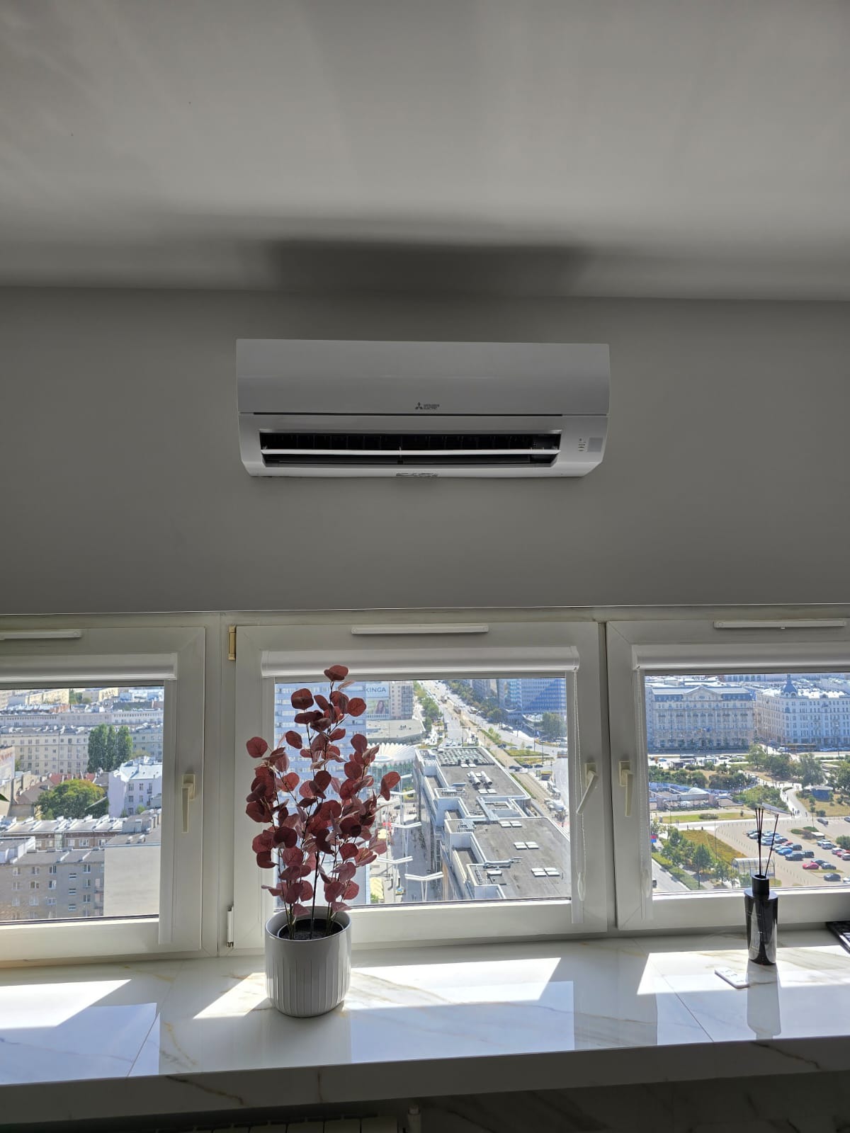 Biały klimatyzator Mitsubishi na ścianie w pokoju z widokiem na miasto przez okno. Na parapecie dekoracje: roślina w doniczce i dyfuzor zapachowy.