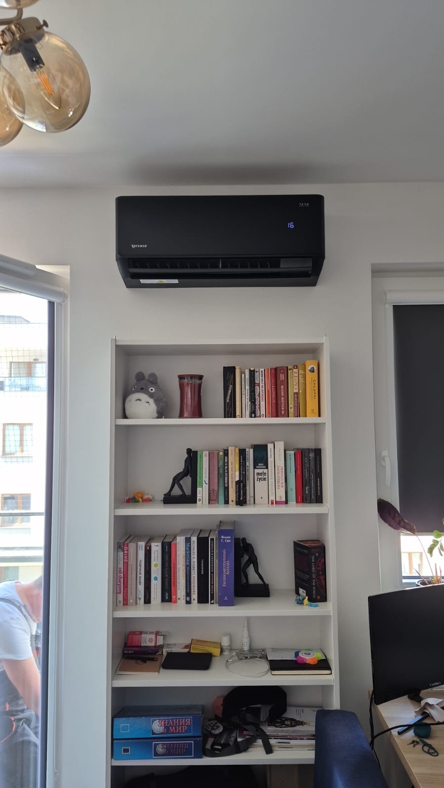 Czarny klimatyzator Rotenso zamontowany nad białym regałem z książkami w jasnym wnętrzu. Widoczny wyświetlacz temperatury i fragment okna.