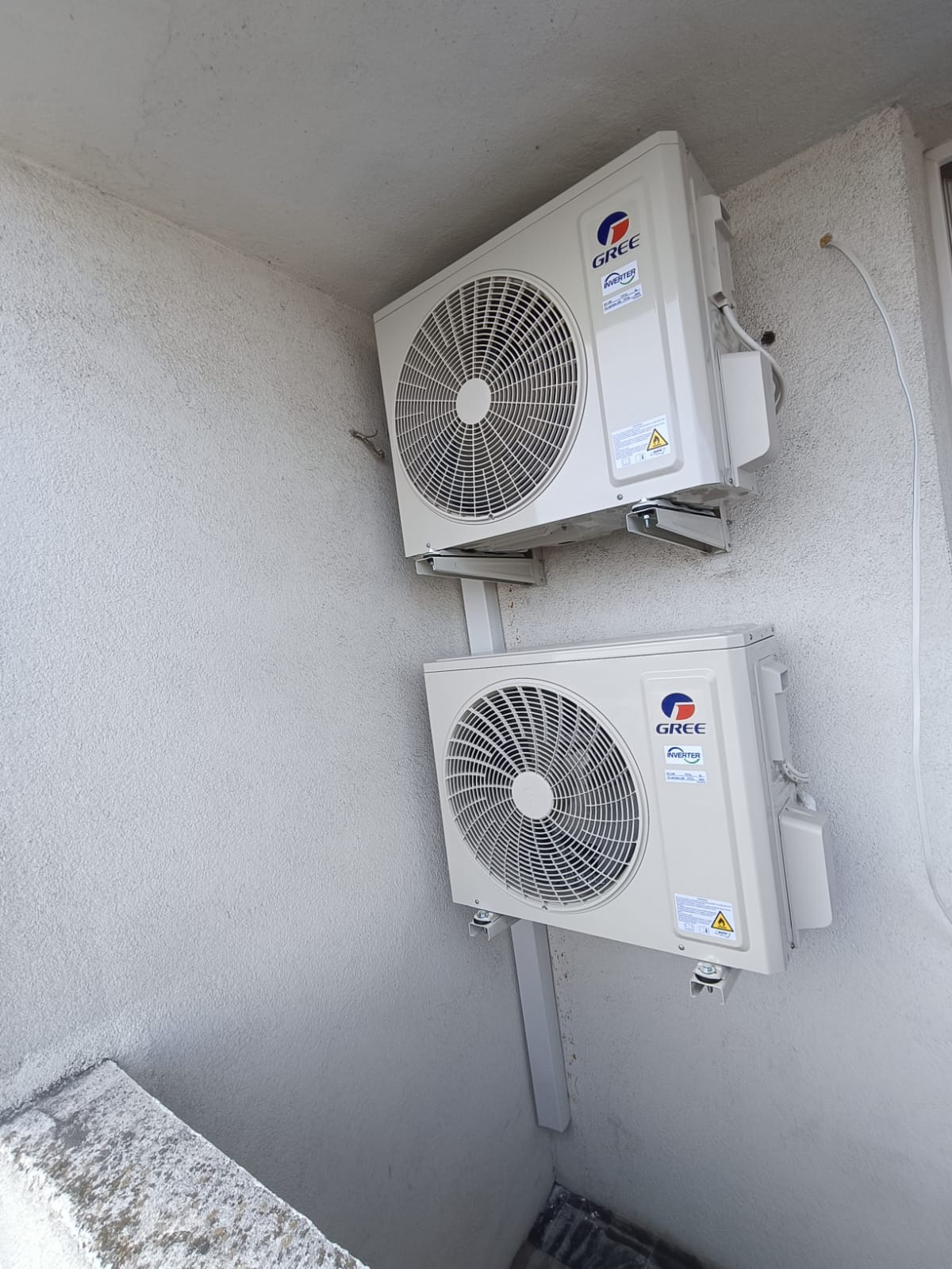 Dwa białe klimatyzatory GREE zamontowane jeden nad drugim na szarej, teksturowanej ścianie zewnętrznej budynku. Widoczne wsporniki montażowe i fragment okna.