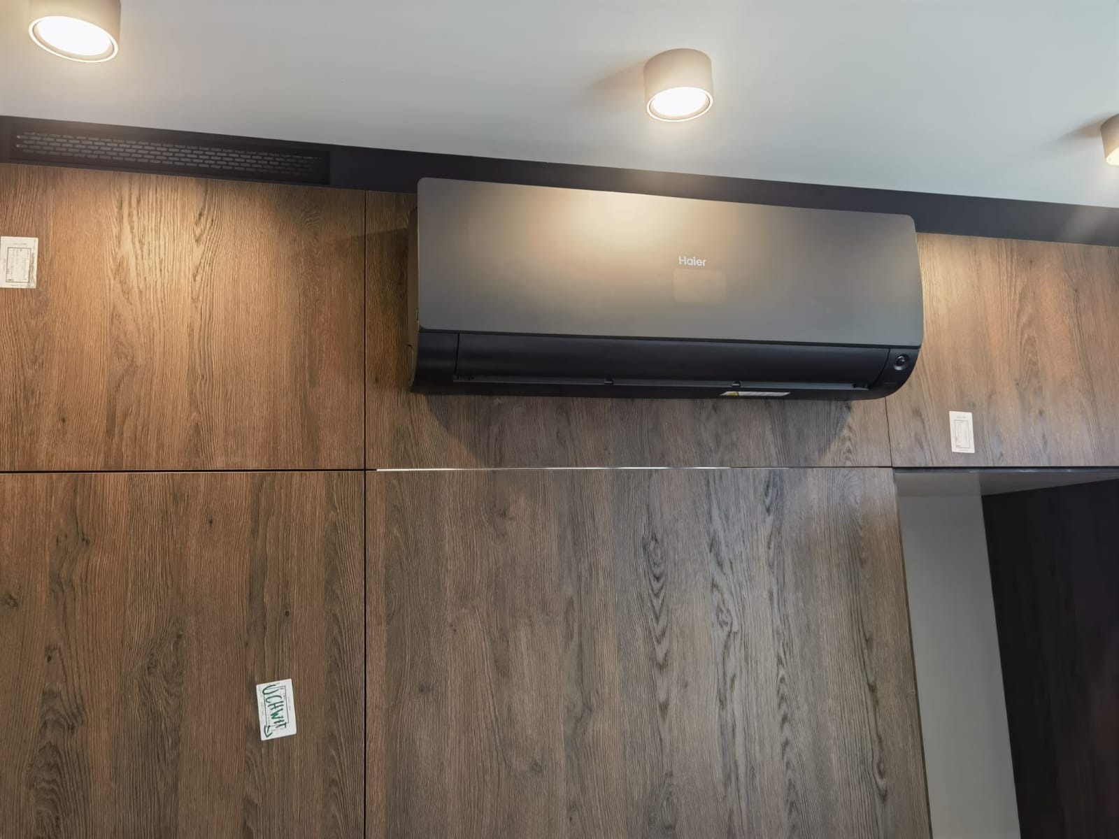 Czarny klimatyzator Haier zamontowany na ścianie z drewnianą okładziną, z widocznymi elementami instalacji i oświetleniem sufitowym.