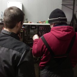 ZUEA BIAŁOWSKI - Dwóch mężczyzn w trakcie pracy przy otwartej szafie sterowniczej z widocznymi przewodami i elementami elektrycznymi. Jeden z nich w czerwonej bluzie manipuluje przy kablach.