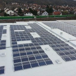 ZUEA BIAŁOWSKI - Rozległa instalacja fotowoltaiczna na białym dachu budynku przemysłowego, z widokiem na miasto i linię kolejową w tle. Panele ułożone w rzędach, częściowo zacienione.