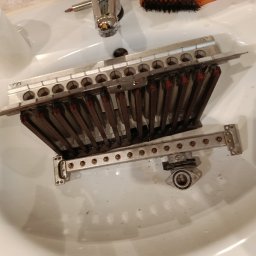 GasTechExpert Mariusz Filipiak - Wymontowany, zabrudzony palnik pieca gazowego w umywalce, widoczne ślady czyszczenia. Obok narzędzia i papier toaletowy. Detal z serwisu pieca gazowego.