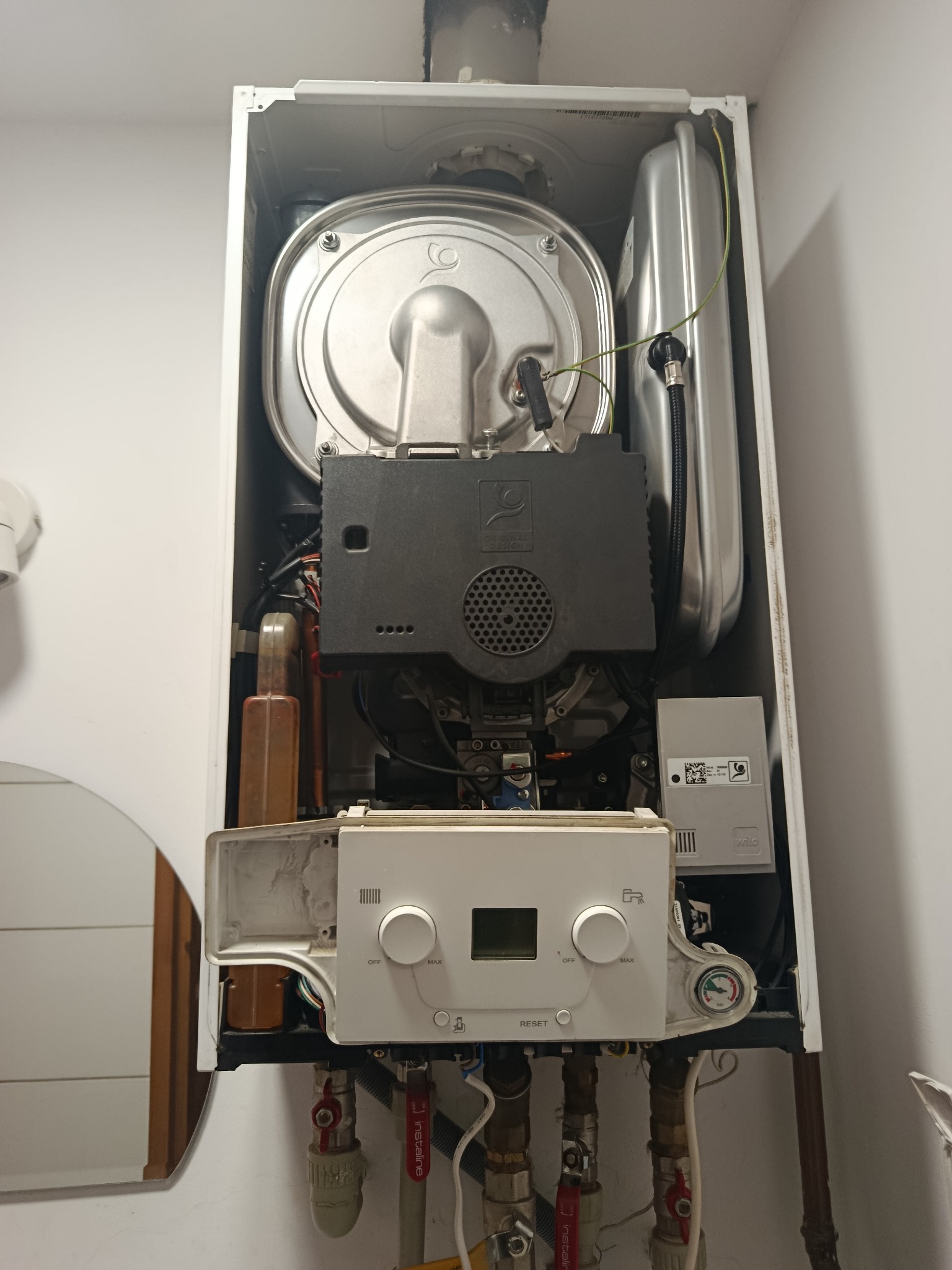 Otwarty piec gazowy z widocznymi elementami wewnętrznymi, w tym palnikiem, elektroniką i rurami. Widoczny manometr i panel sterowania.