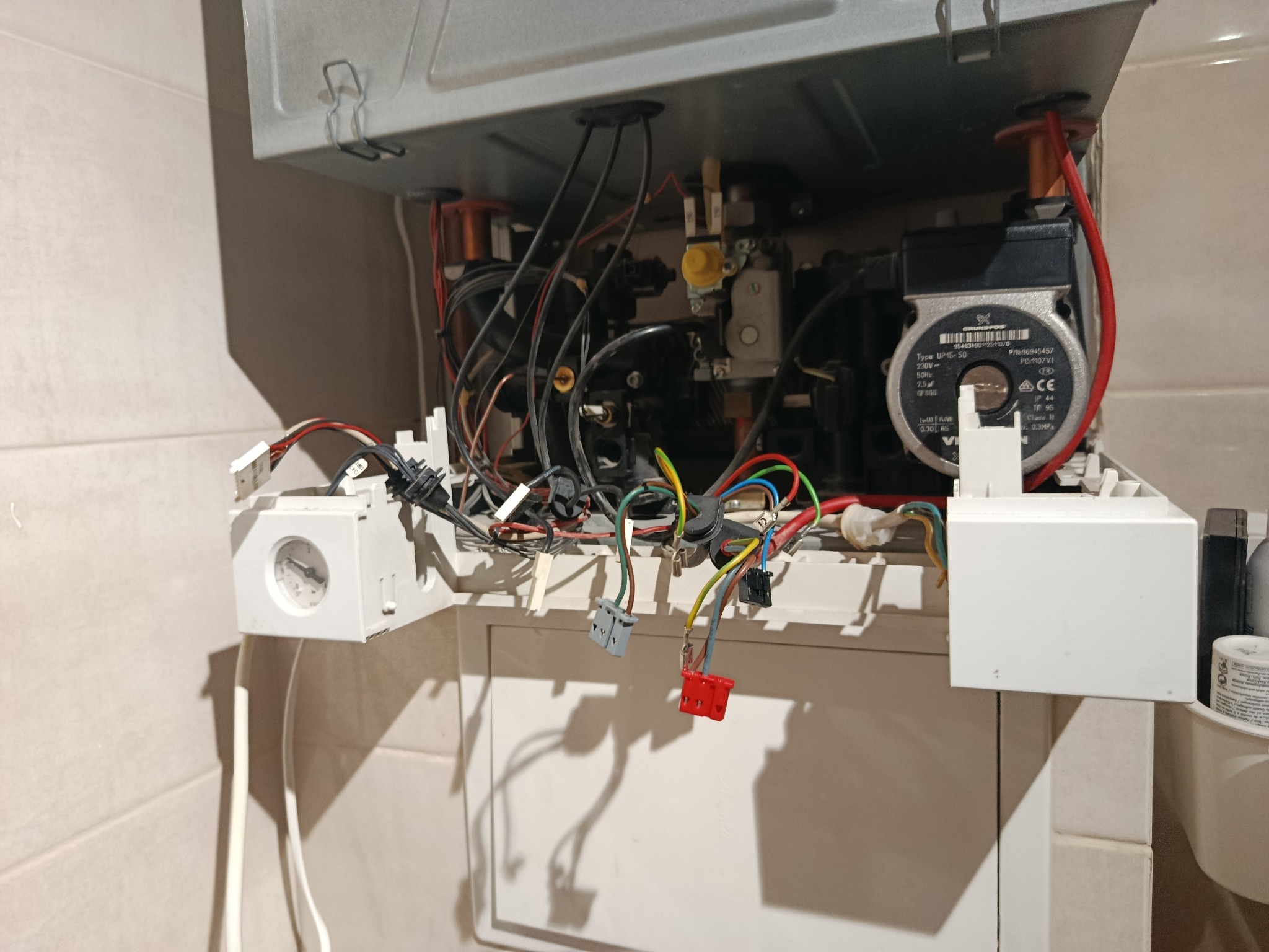 Otwarty piec gazowy z widocznymi przewodami, elementami instalacji i pompą obiegową. Widoczne złącza elektryczne i manometr. Ujęcie z bliska, wnętrze urządzenia.