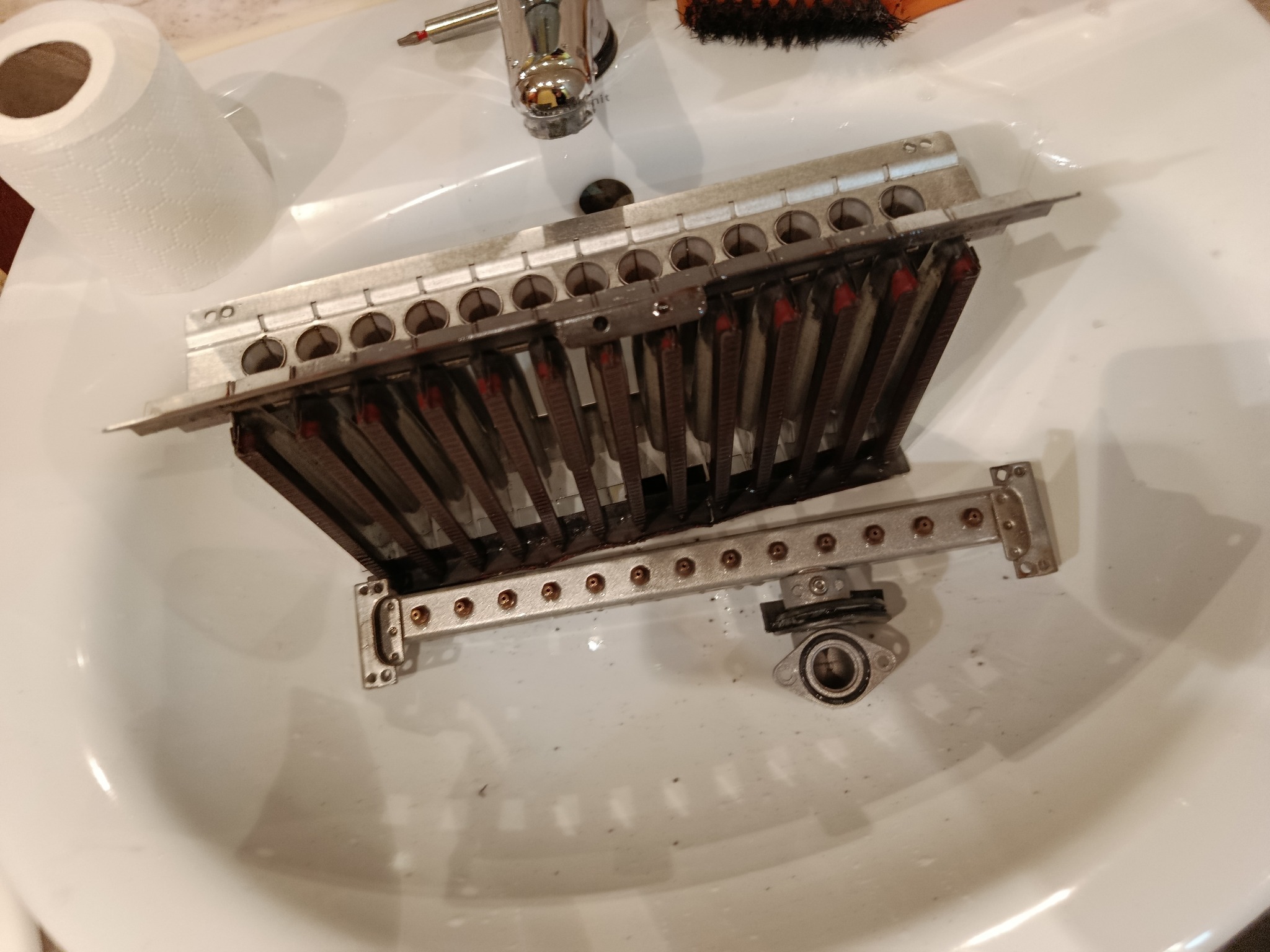 Wymontowany, zabrudzony palnik pieca gazowego w umywalce, widoczne ślady czyszczenia. Obok narzędzia i papier toaletowy. Detal z serwisu pieca gazowego.