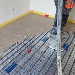 UdelemeTo s.r.o. - Wylewka samopoziomująca częściowo zakrywająca siatkę grzewczą podłogową z rurkami, widoczne narzędzia i fragment nogi pracownika w odzieży roboczej.