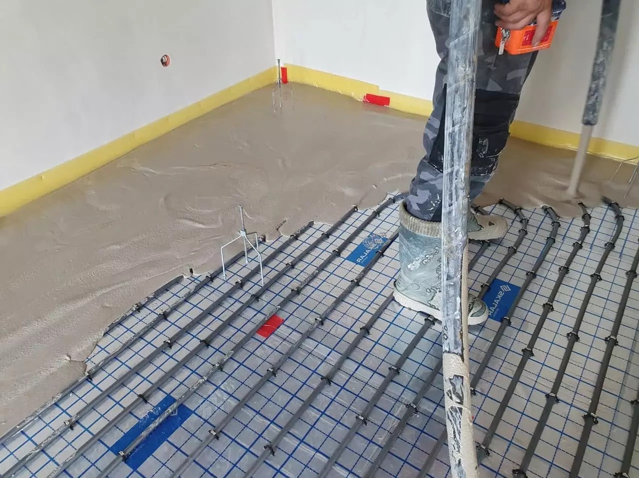 Wylewka samopoziomująca częściowo zakrywająca siatkę grzewczą podłogową z rurkami, widoczne narzędzia i fragment nogi pracownika w odzieży roboczej.