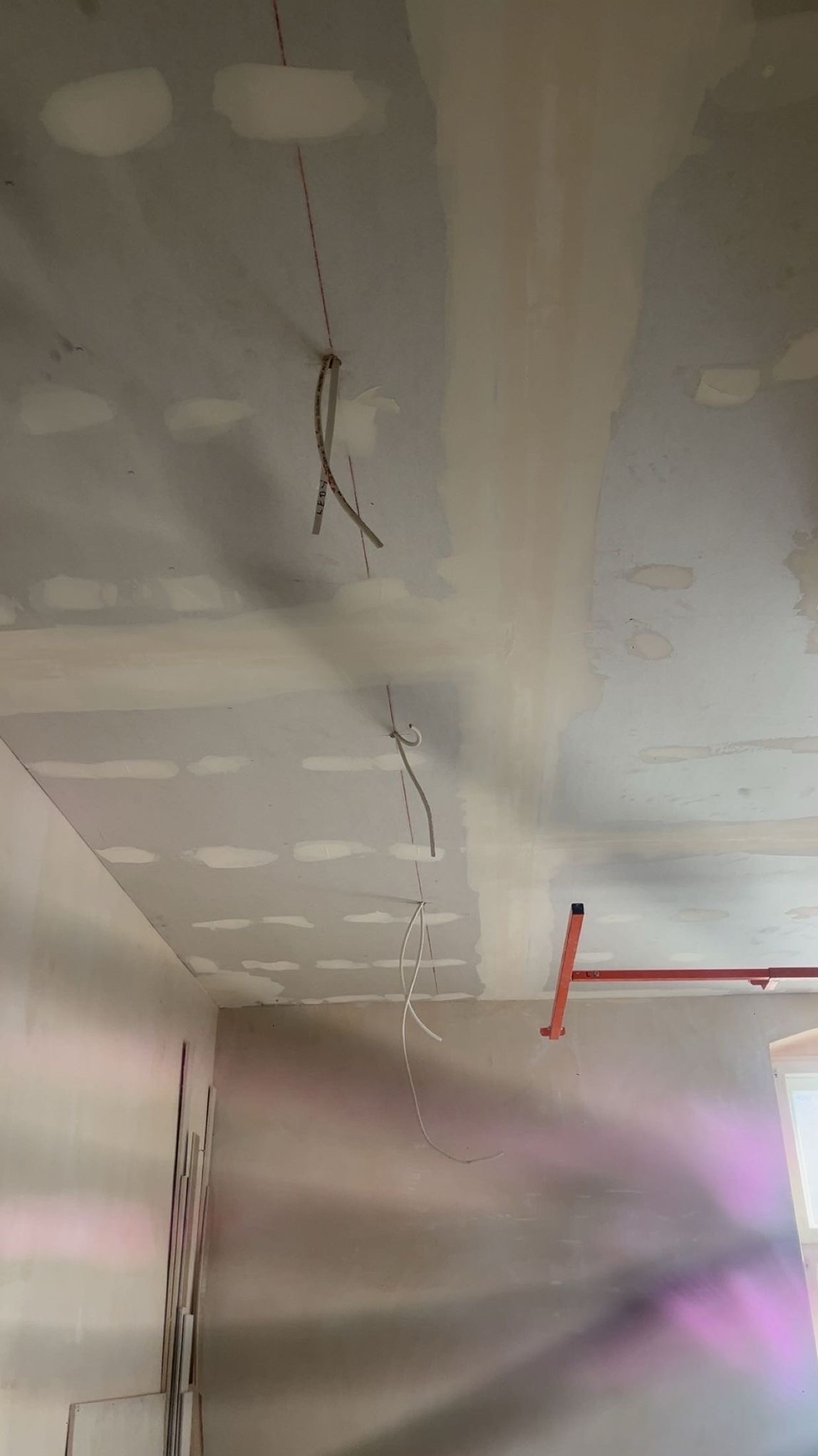 Sufit z płyt kartonowo-gipsowych w trakcie montażu, widoczne kable i szpachlowane łączenia. Fragment ściany z widocznym oknem i element rusztowania.