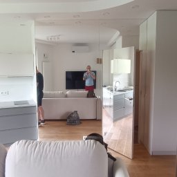 Taras Bulba Company - Jasne wnętrze po malowaniu: salon z sofą i listwami przypodłogowymi, widok na kuchnię w lustrzanej ścianie. Osoba robiąca zdjęcie widoczna w odbiciu.
