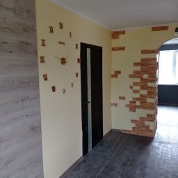 Taras Bulba Company - Wykończone wnętrze: kremowe ściany z dekoracyjnym zegarem i imitacją cegły, ciemna stolarka drzwiowa, panele podłogowe w odcieniach szarości i beżu. Widok na kolejne pomieszczenie.