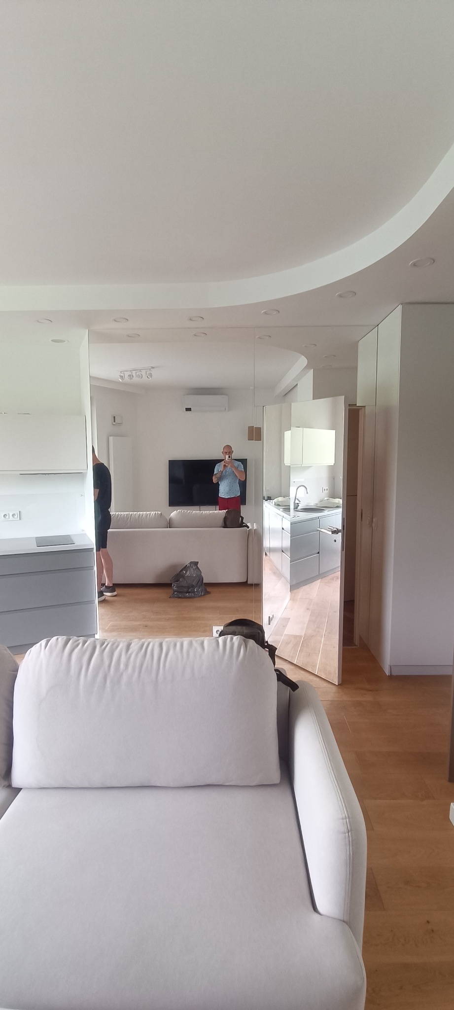 Jasne wnętrze po malowaniu: salon z sofą i listwami przypodłogowymi, widok na kuchnię w lustrzanej ścianie. Osoba robiąca zdjęcie widoczna w odbiciu.