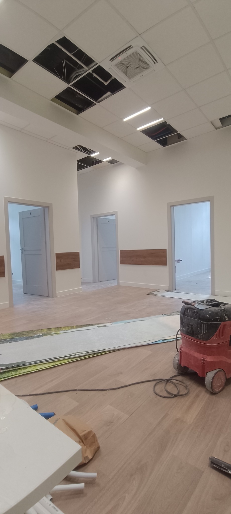 Wnętrze biura po malowaniu ścian na biało, z widocznym odkurzaczem przemysłowym i elementami sufitu podwieszanego w trakcie montażu. Widoczne trzy drzwi.