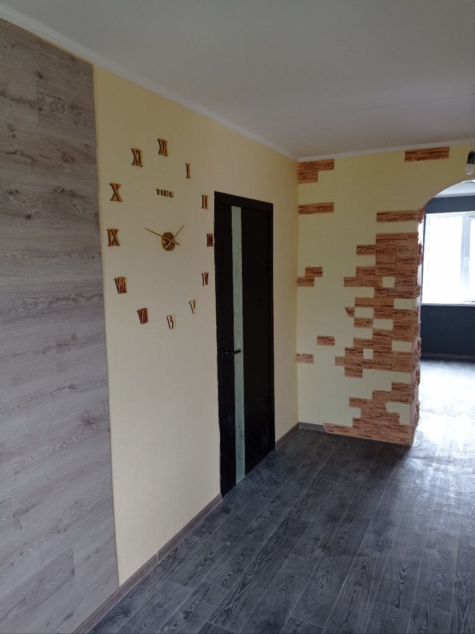 Wykończone wnętrze: kremowe ściany z dekoracyjnym zegarem i imitacją cegły, ciemna stolarka drzwiowa, panele podłogowe w odcieniach szarości i beżu. Widok na kolejne pomieszczenie.