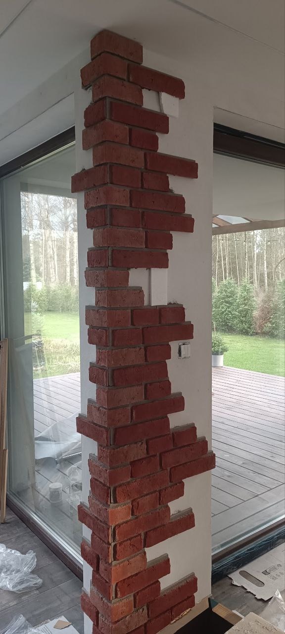 Częściowo wykończona ściana z dekoracyjnej cegły wewnątrz jasnego pomieszczenia z dużym oknem. Widoczne niedokończone fragmenty i materiały budowlane.