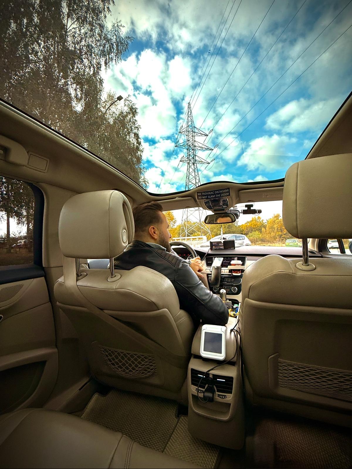 Wnętrze beżowego auta z panoramicznym dachem, widok na kierowcę w skórzanej kurtce i słup wysokiego napięcia na tle błękitnego nieba z chmurami.