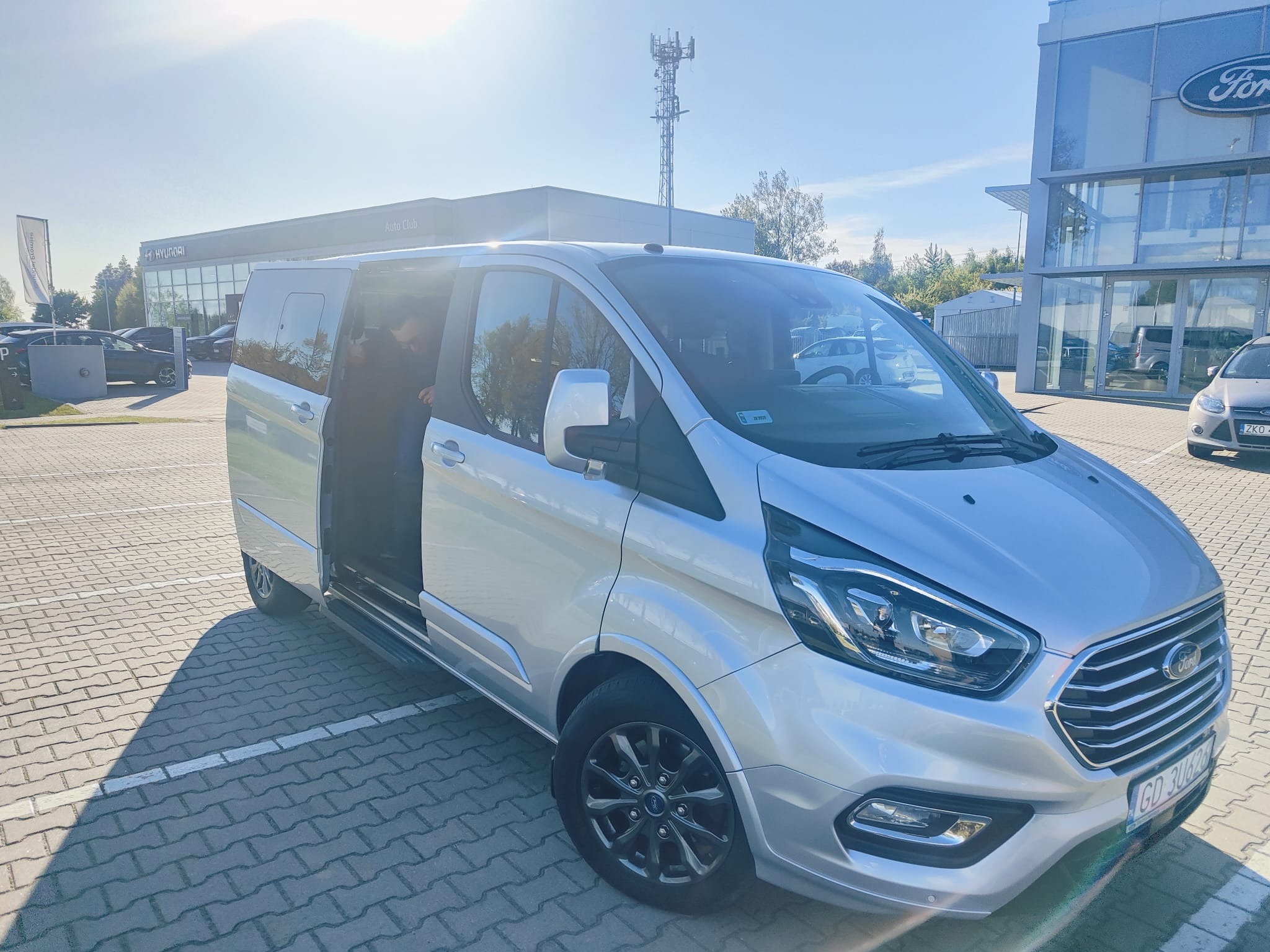 Srebrny Ford Transit Custom z otwartymi drzwiami przesuwnymi na brukowanym parkingu, w tle salon samochodowy i wieża telekomunikacyjna w słoneczny dzień.