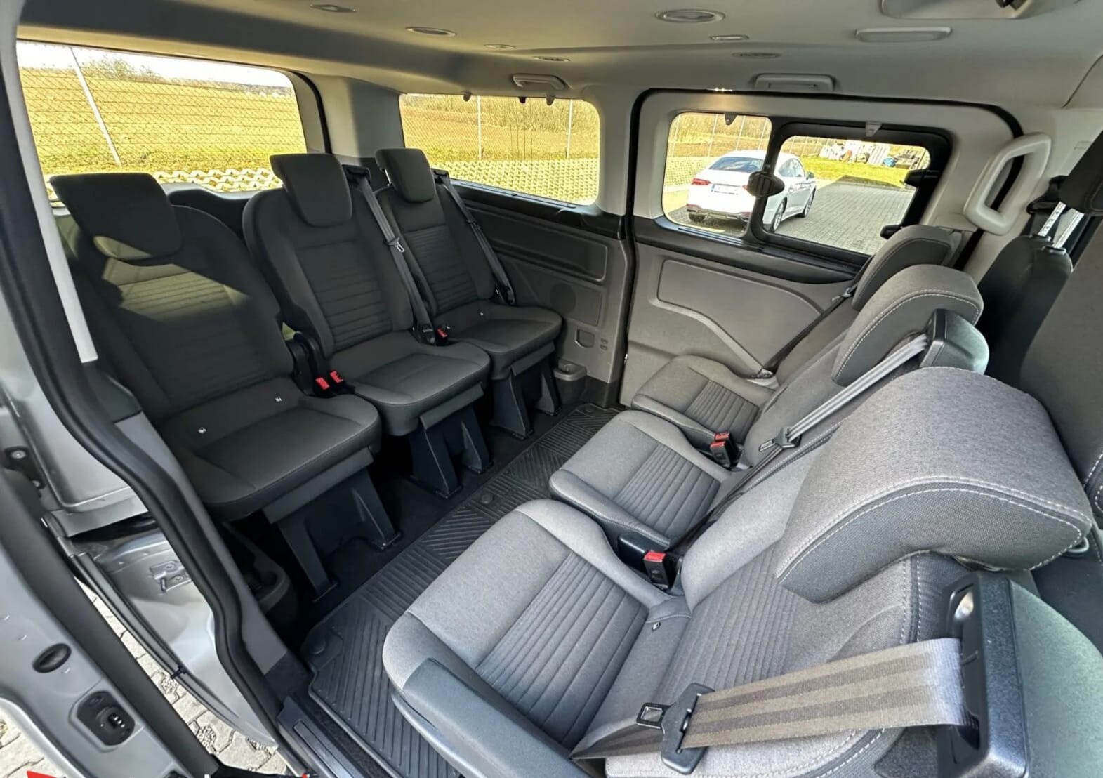 Wnętrze przestronnego busa z trzema rzędami siedzeń w odcieniach szarości. Widok z otwartych drzwi na komfortowe fotele z pasami bezpieczeństwa i widokiem na zewnątrz.