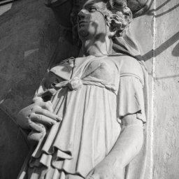 Robert Chmara - Czarno-białe zdjęcie kamiennej rzeźby kobiety w drapowanej szacie, z dzbanami na głowie, umieszczonej na ścianie budynku, oświetlonej z góry.