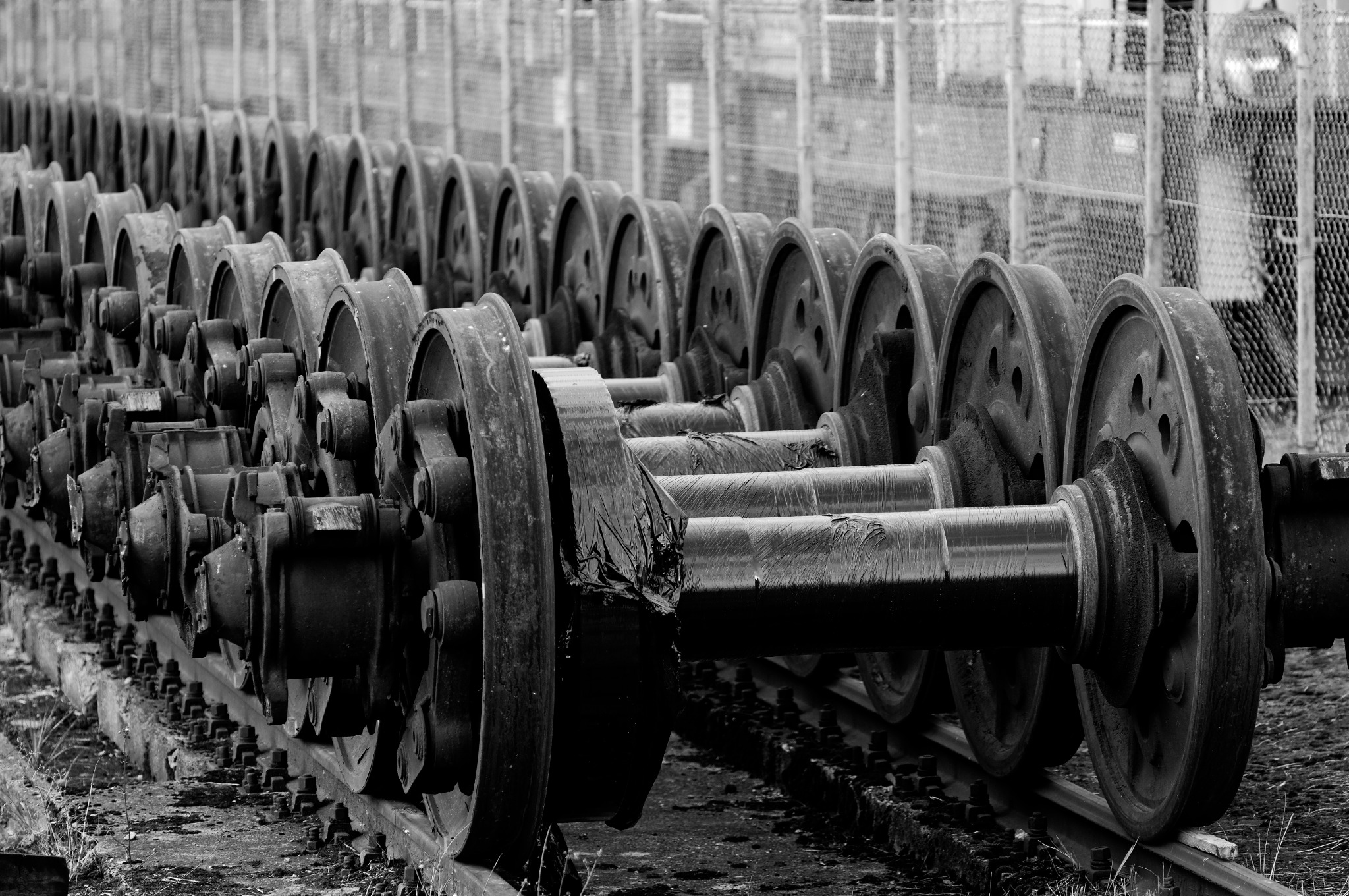 Rząd metalowych kół na szynach w czerni i bieli, z widocznymi śladami rdzy i zużycia, sugerujący opuszczony lub przemysłowy charakter miejsca.